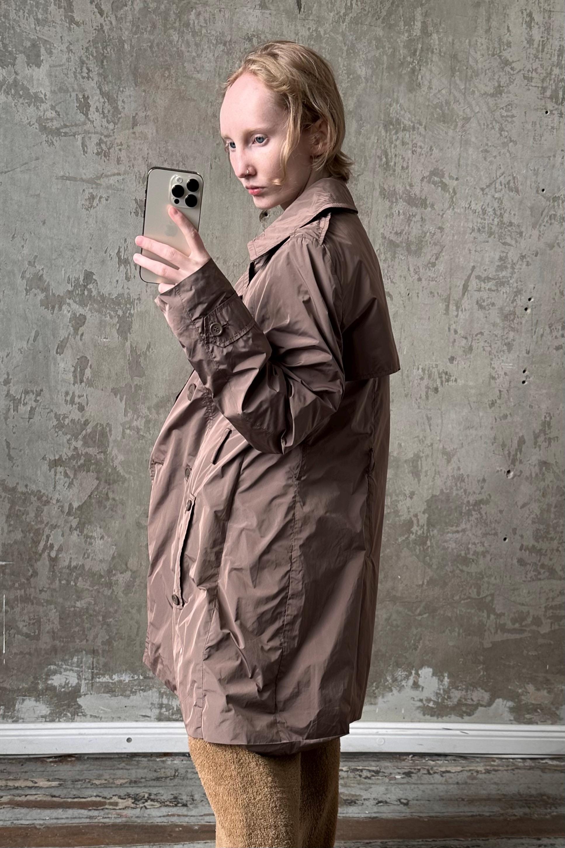 A person in a brown coat is holding a smartphone and taking a selfie in front of a concrete wall.