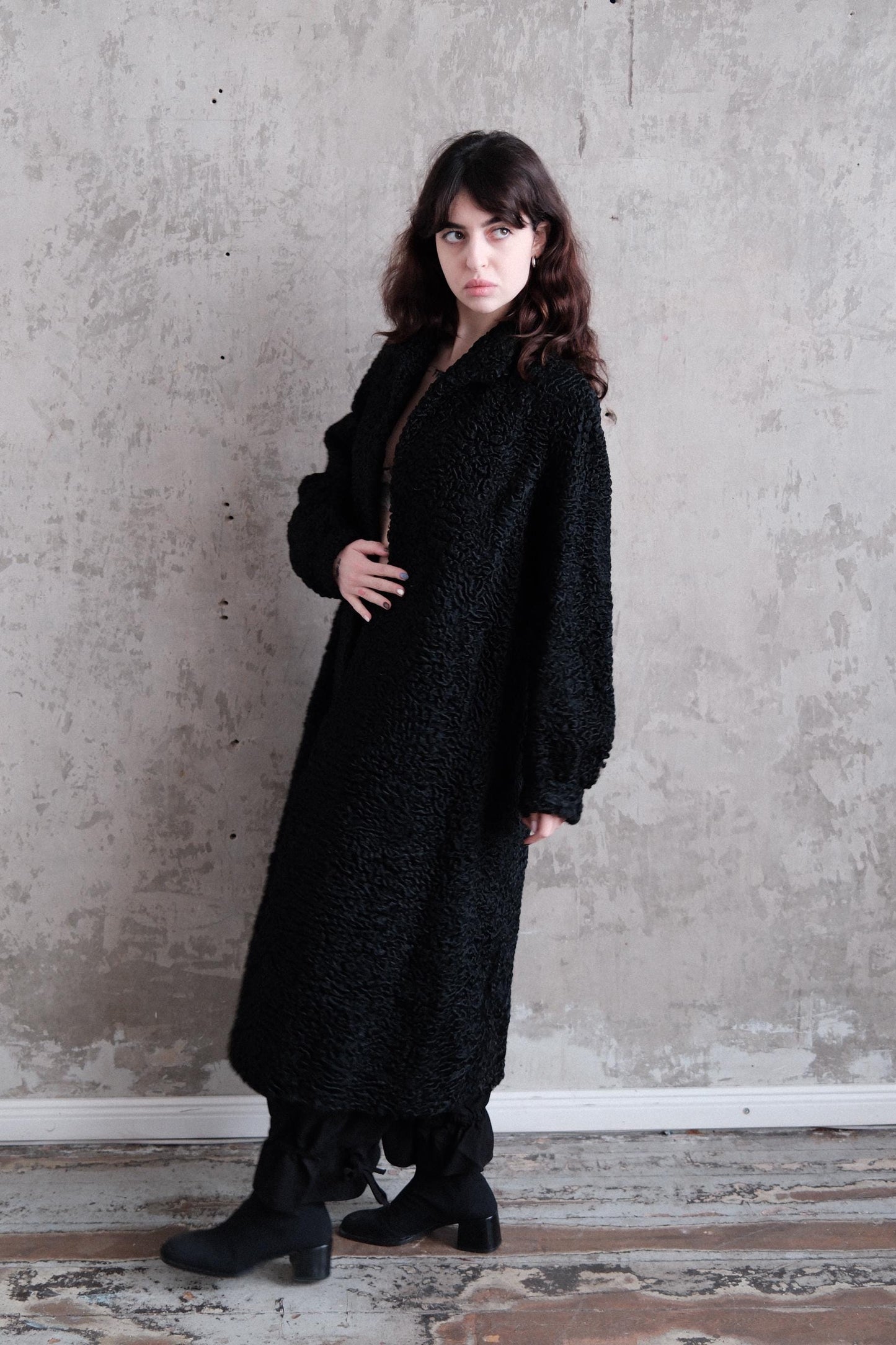 A woman wearing a long black coat stands in a room with a concrete wall.