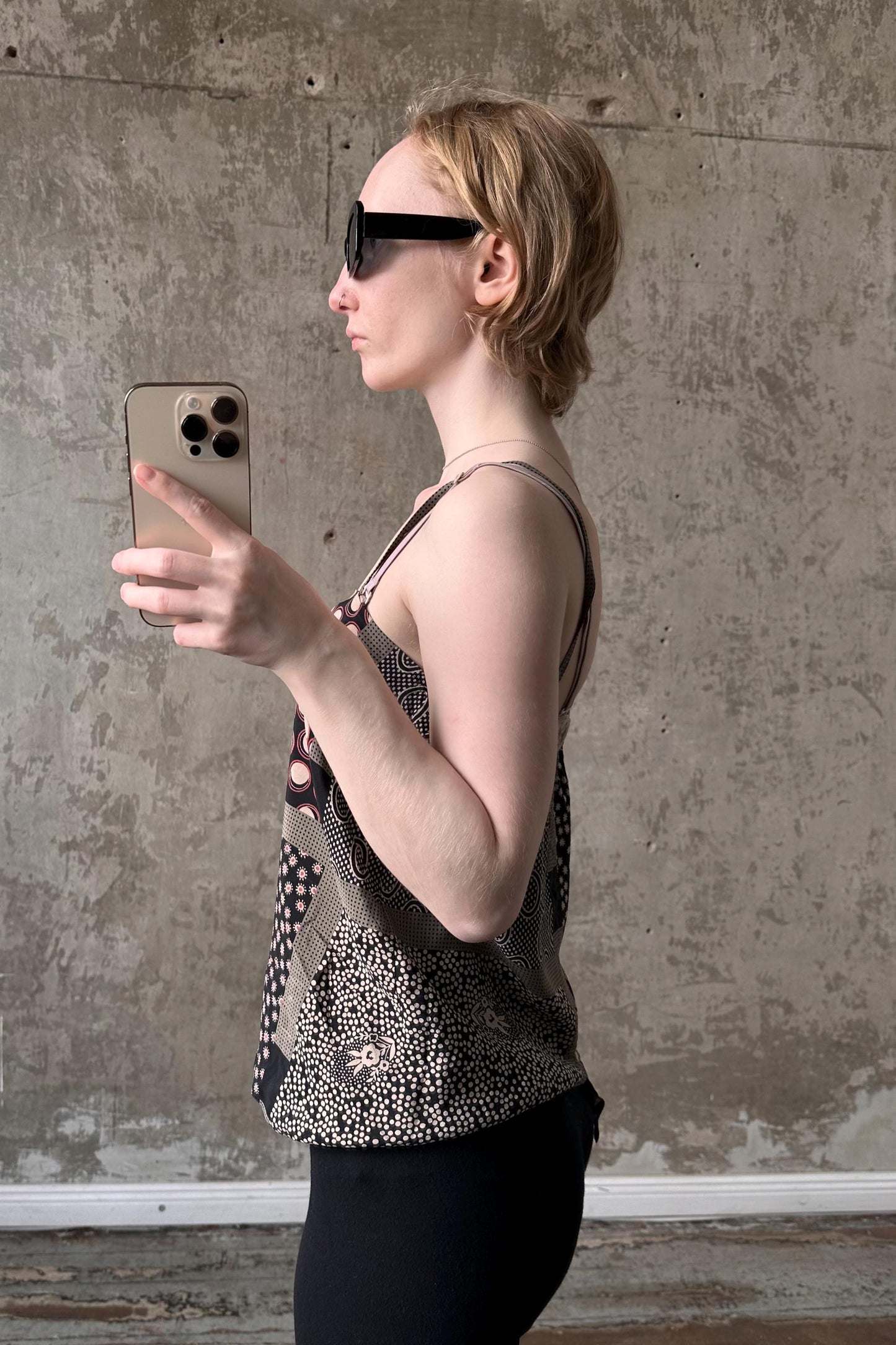 A woman wearing a patterned top and sunglasses is holding a phone and looking at the screen, standing in front of a concrete wall.