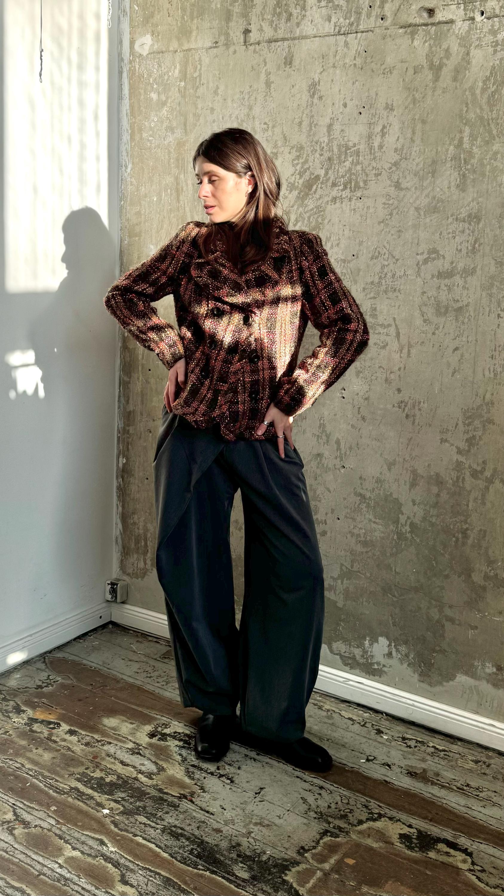 A woman wearing a brown and black patterned jacket stands in a room with a concrete wall and wooden floor.