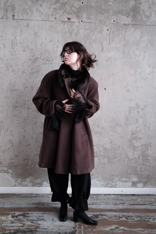 A person wearing a brown coat and black pants stands against a concrete wall, looking up with a serious expression.
