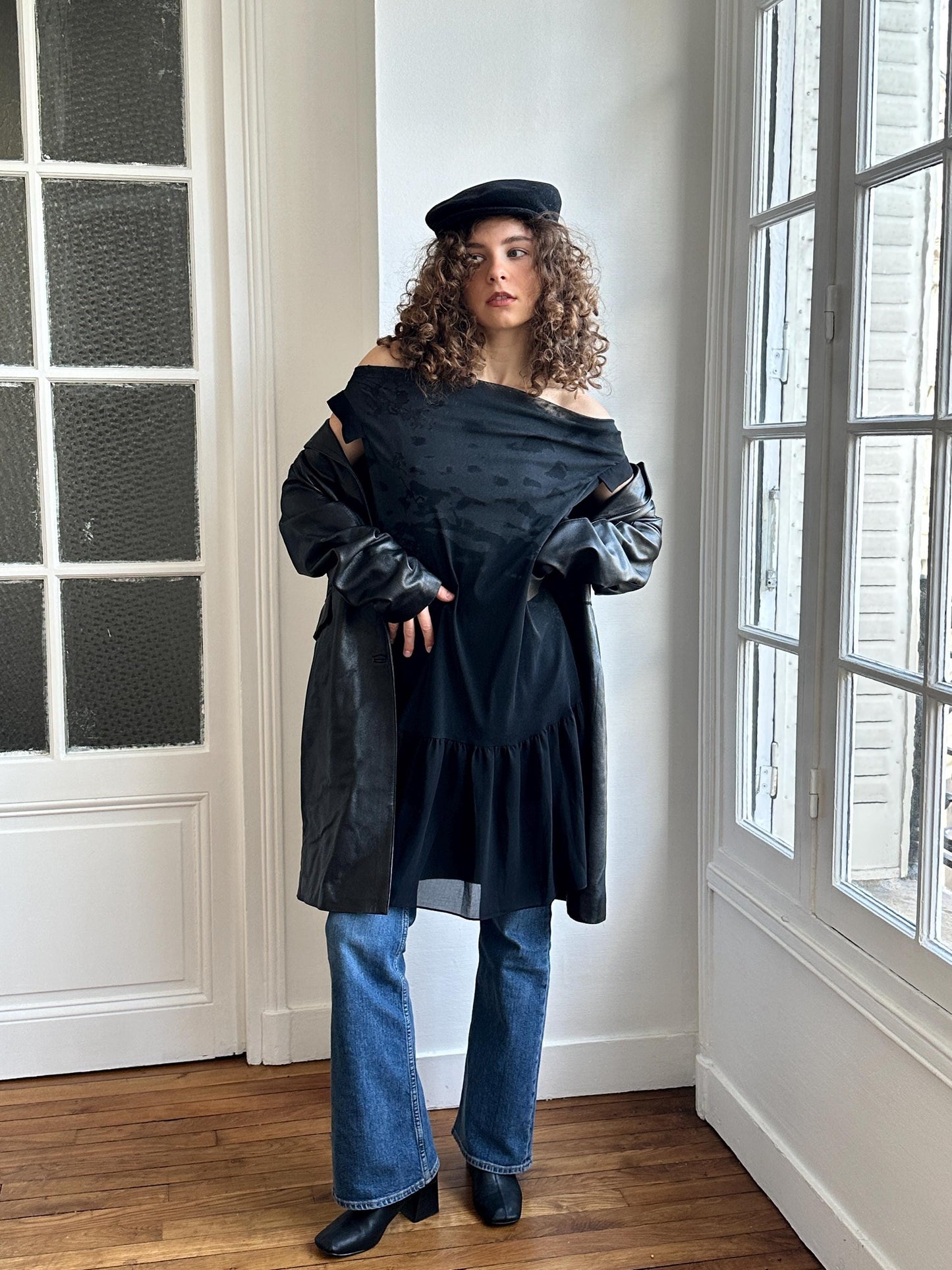 A woman with curly hair wearing a black off-the-shoulder top, a black leather jacket, and blue jeans stands in a room with a window.