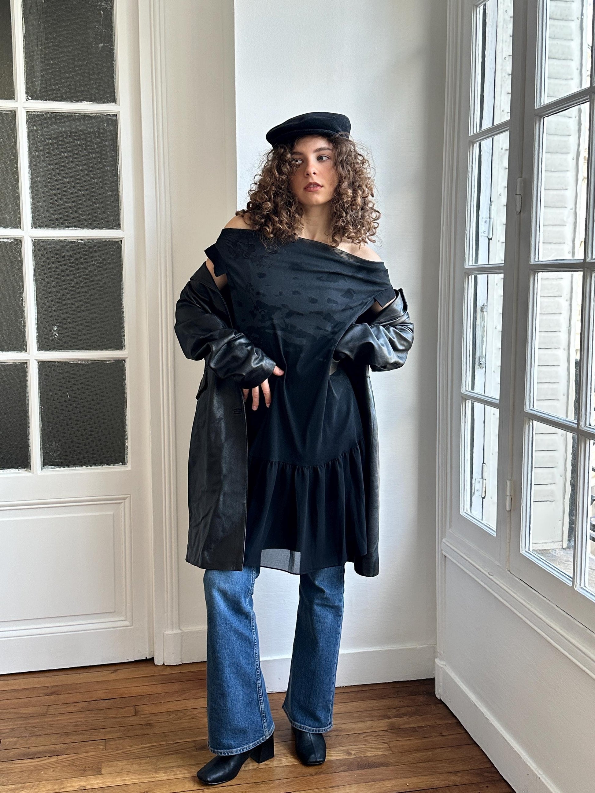 A woman with curly hair wearing a black off-the-shoulder top, a black leather jacket, and blue jeans stands in a room with a window.