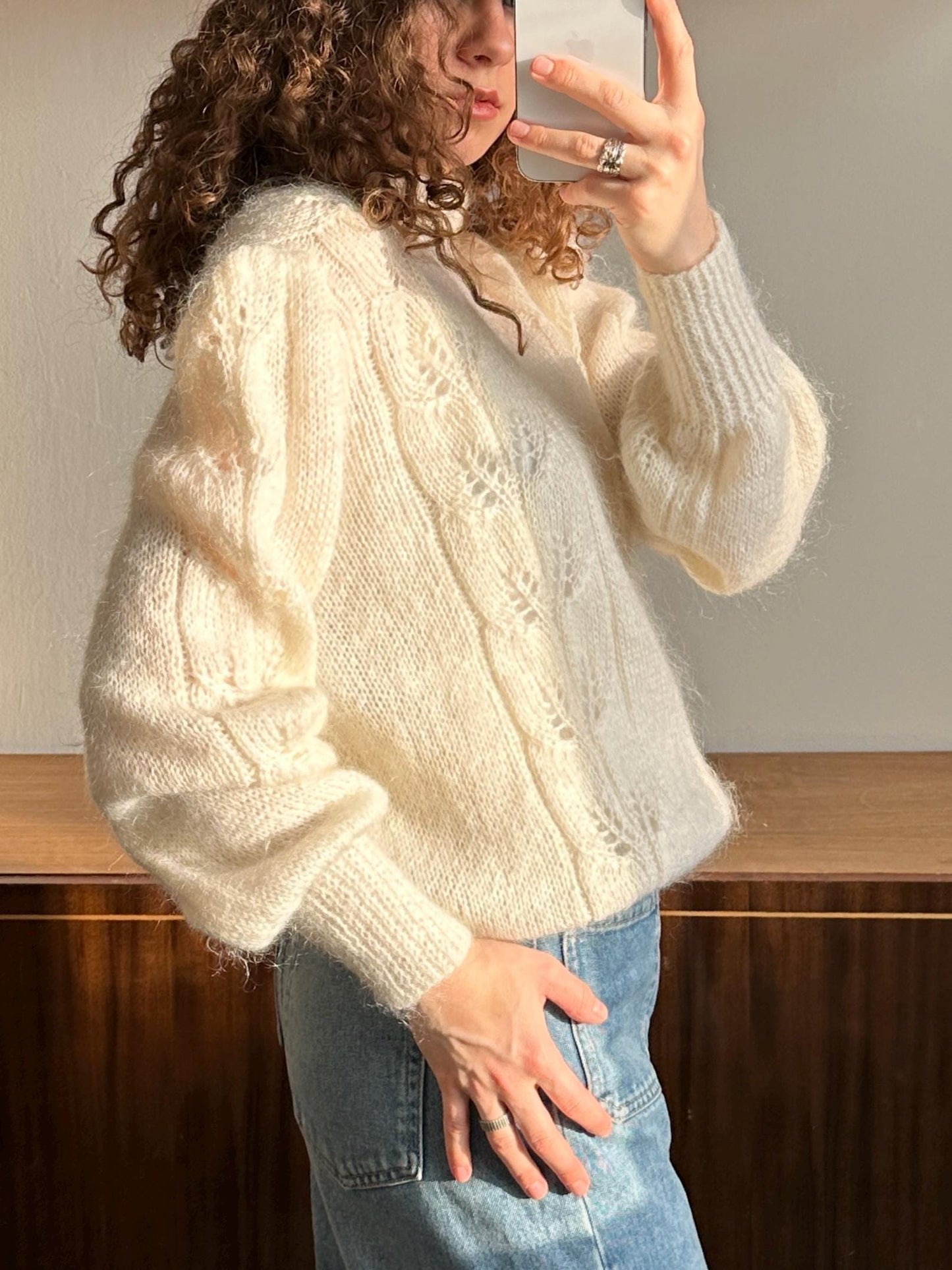 A woman with curly hair is taking a selfie with a white sweater and blue jeans.