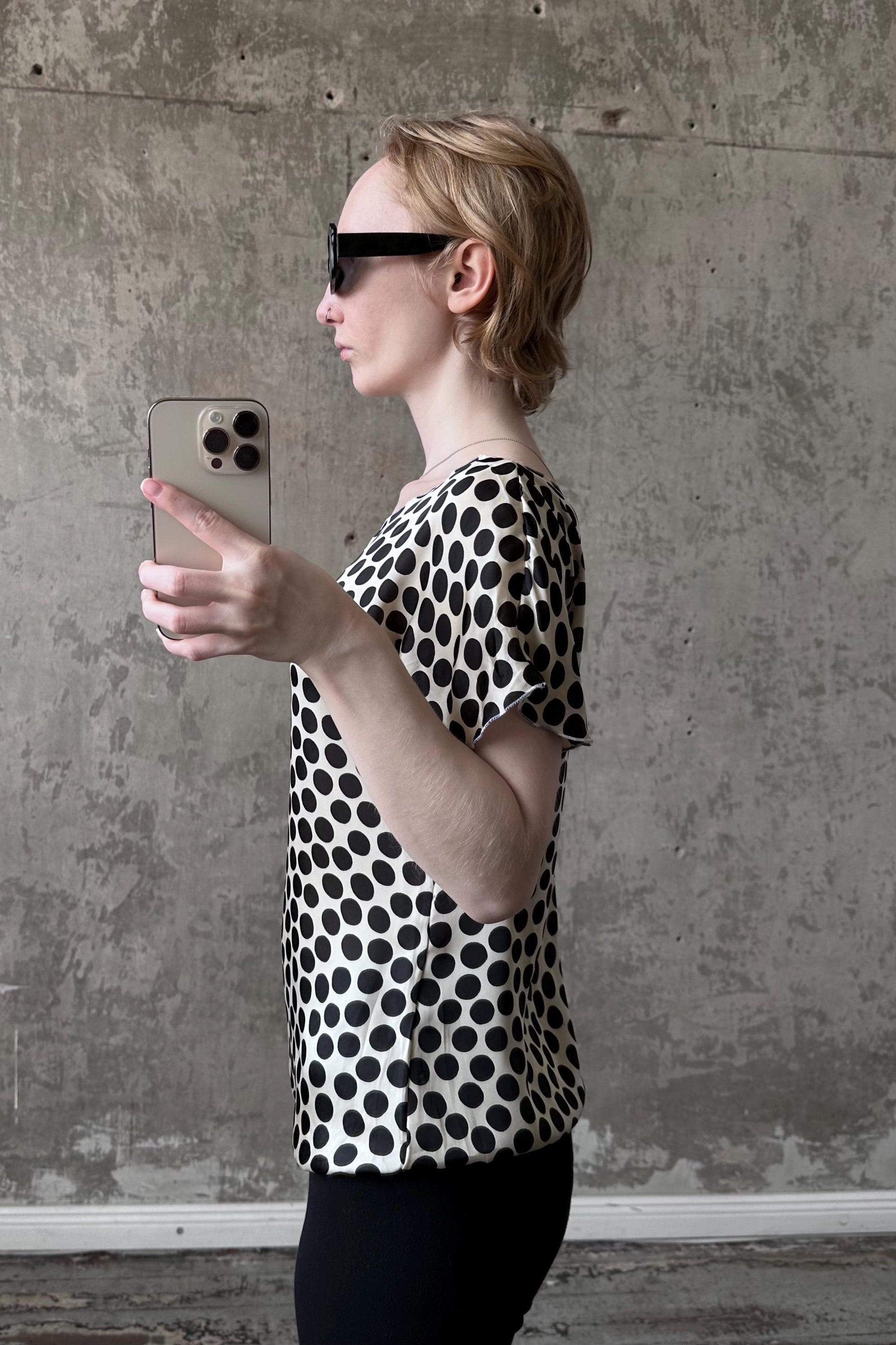 A woman wearing a black and white polka dot blouse is holding a smartphone and taking a selfie in front of a concrete wall.