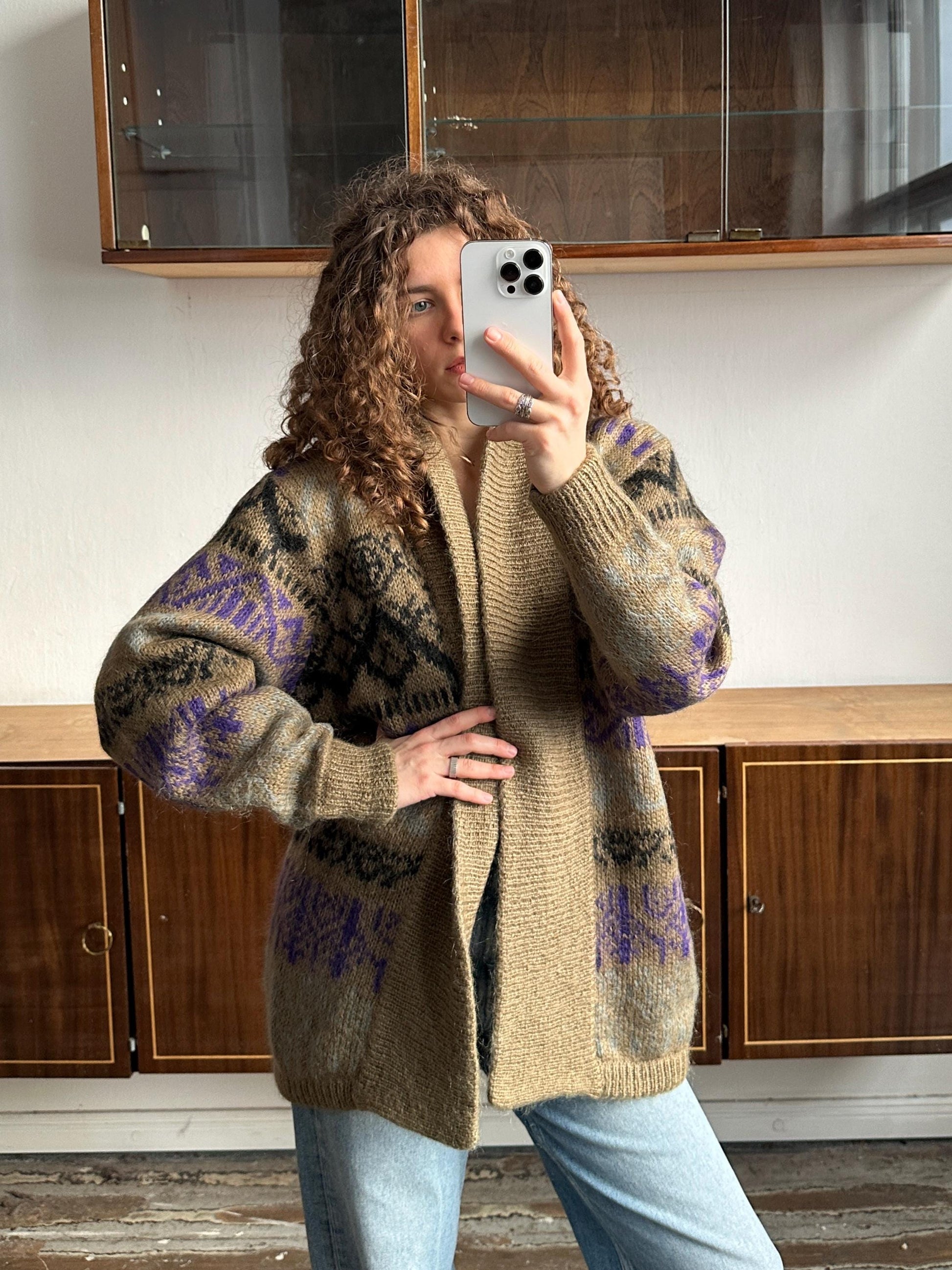 A woman with curly hair is taking a selfie with a smartphone, wearing a beige cardigan with purple and black designs.