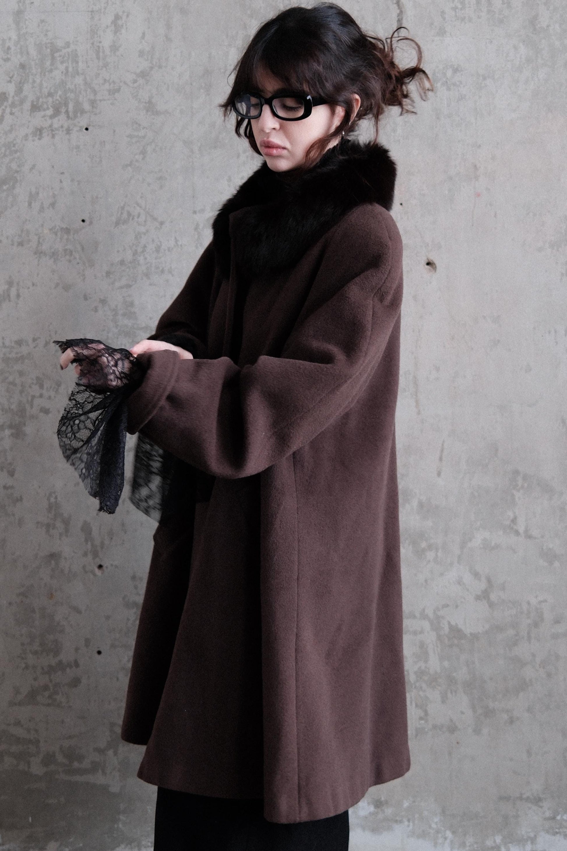 A woman wearing a brown coat and glasses is standing against a concrete wall.