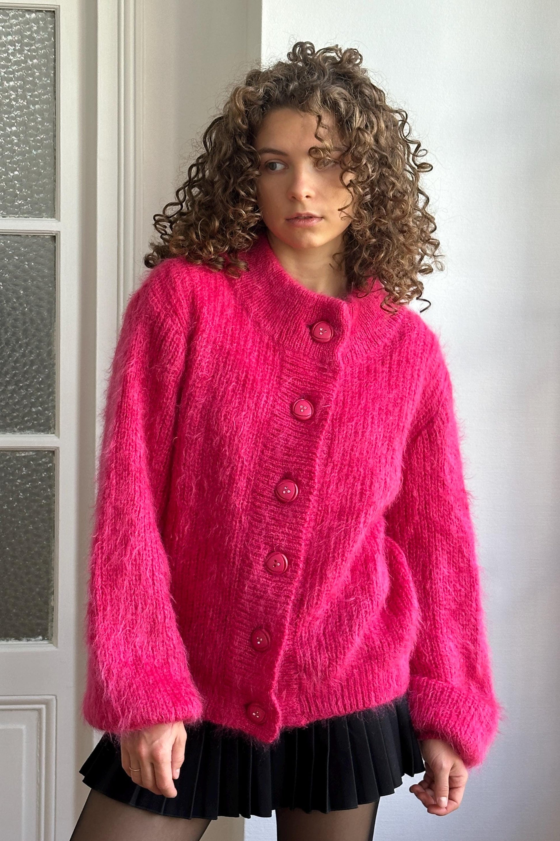 A woman with curly hair wearing a bright pink cardigan and black skirt stands in front of a white door.