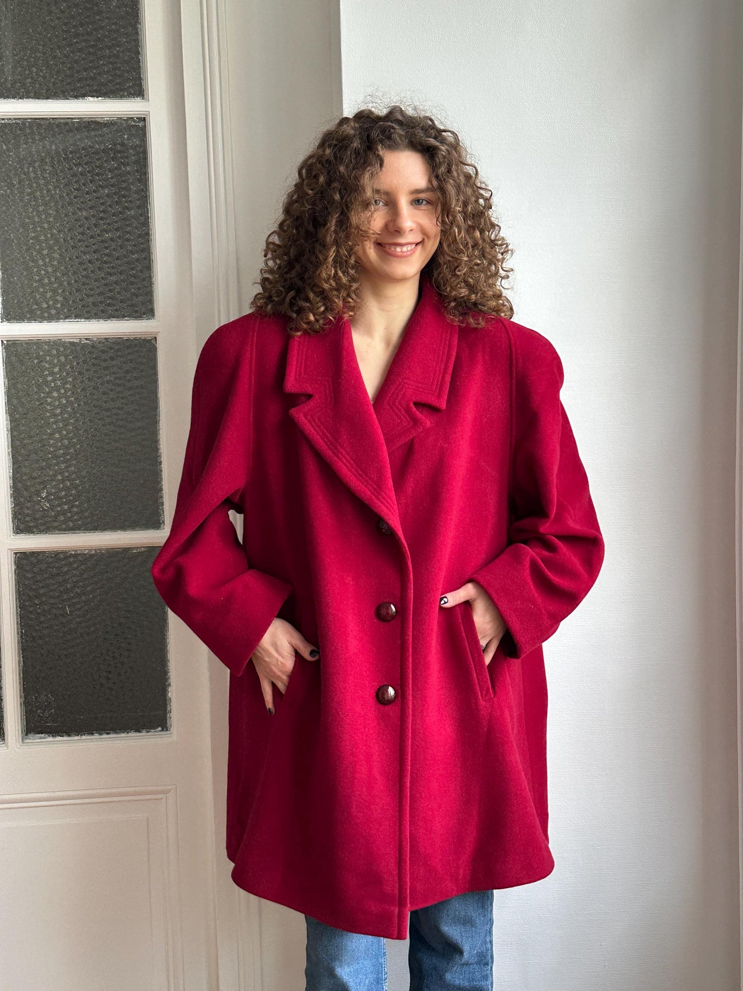 A person wearing a red coat is smiling and leaning against a white wall.