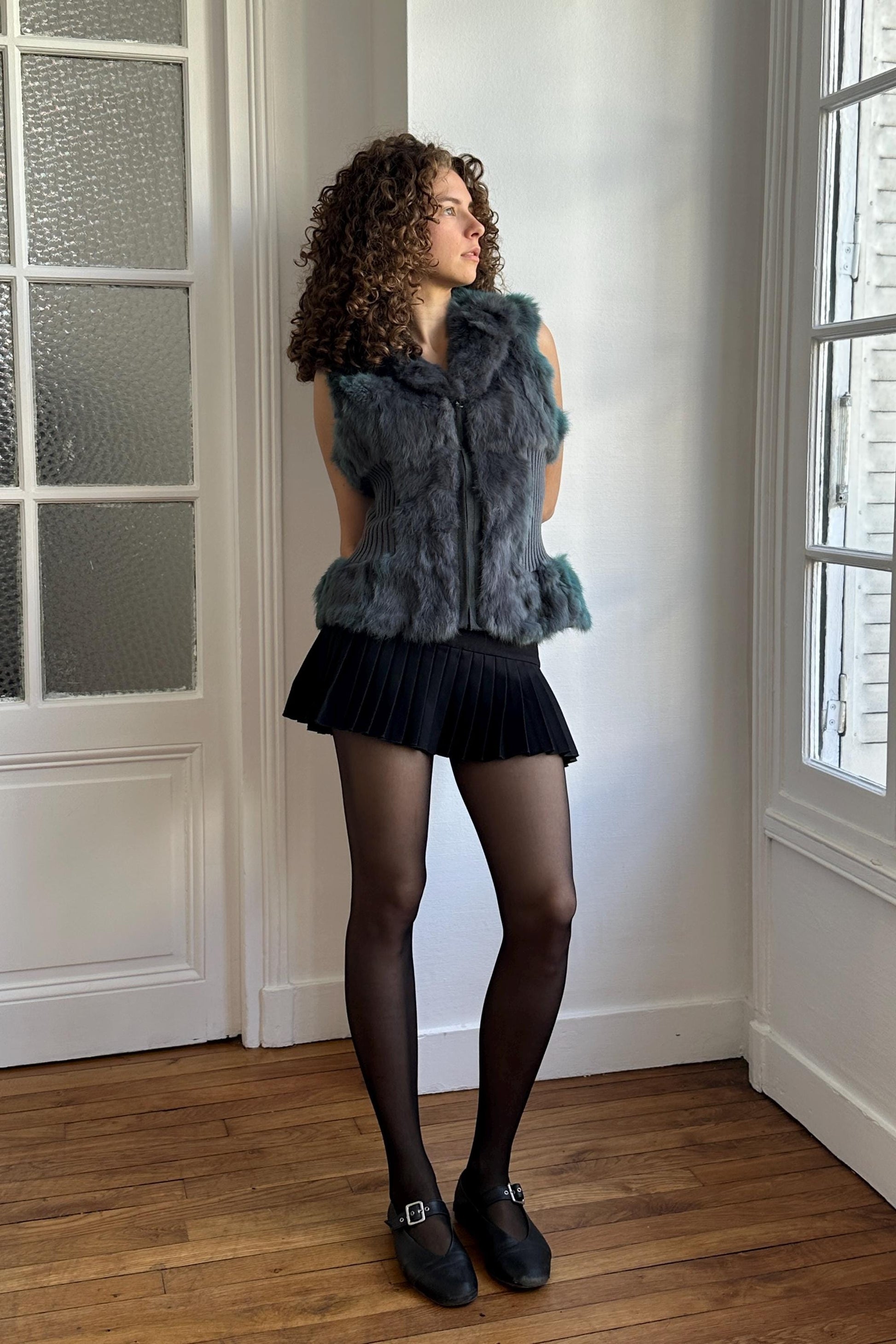 A woman with curly hair stands in a room, wearing a gray fur vest, black skirt, and black shoes.