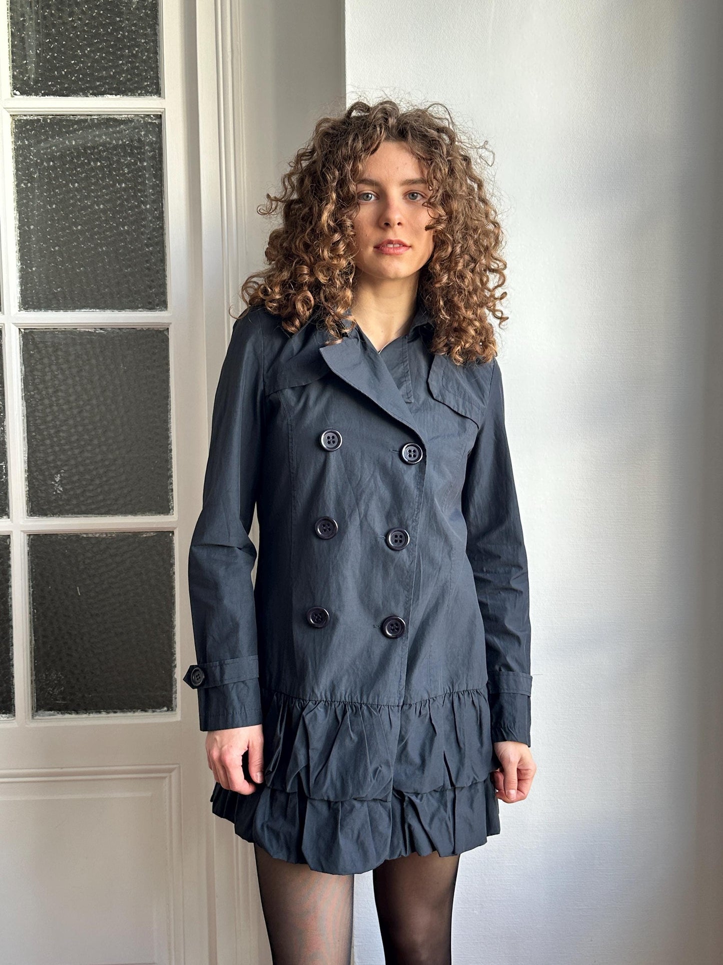 A woman with curly hair wearing a gray coat and skirt is standing in front of a window.