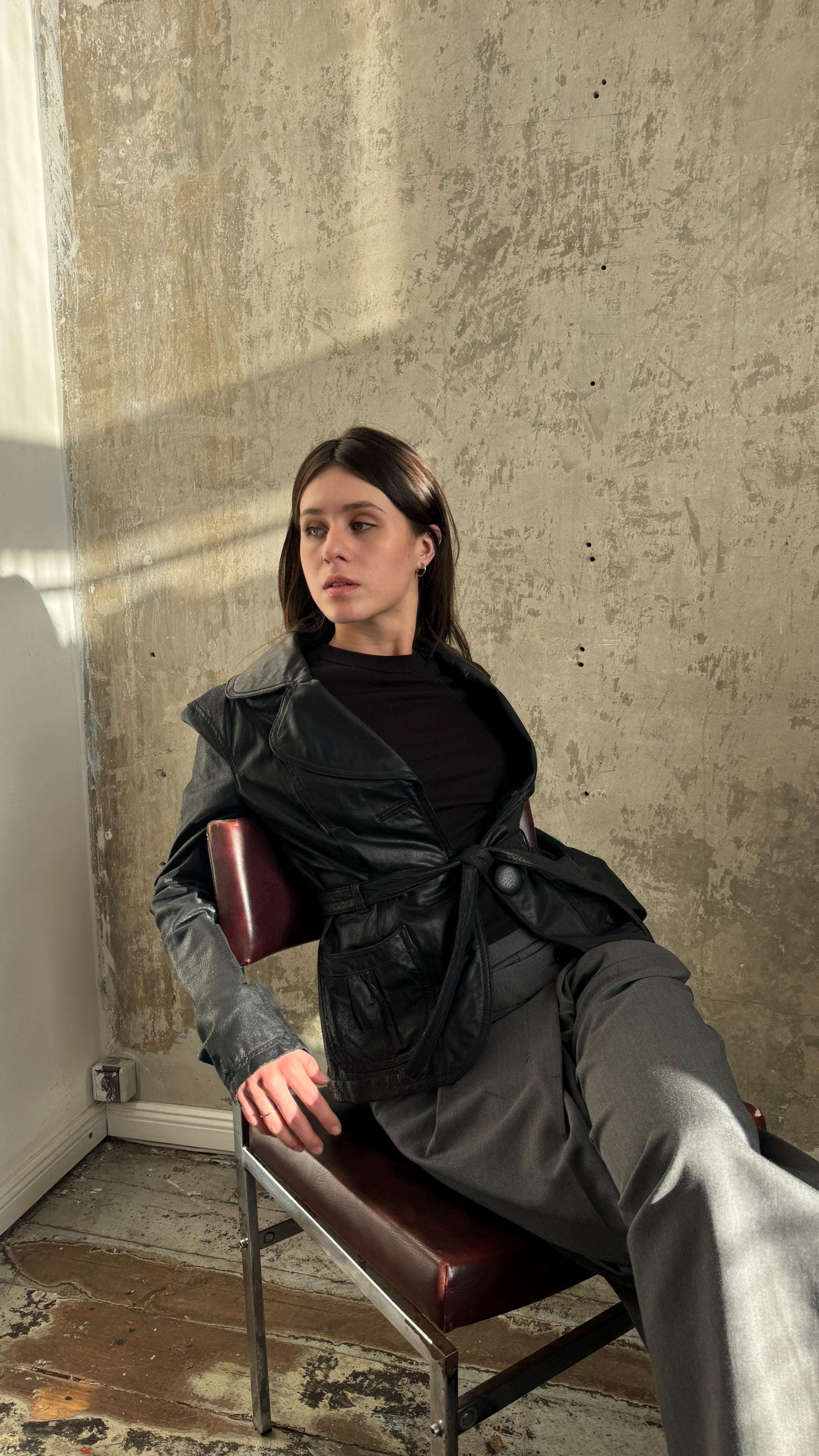 A woman in a black jacket sits on a chair, looking off to the side with a serious expression.