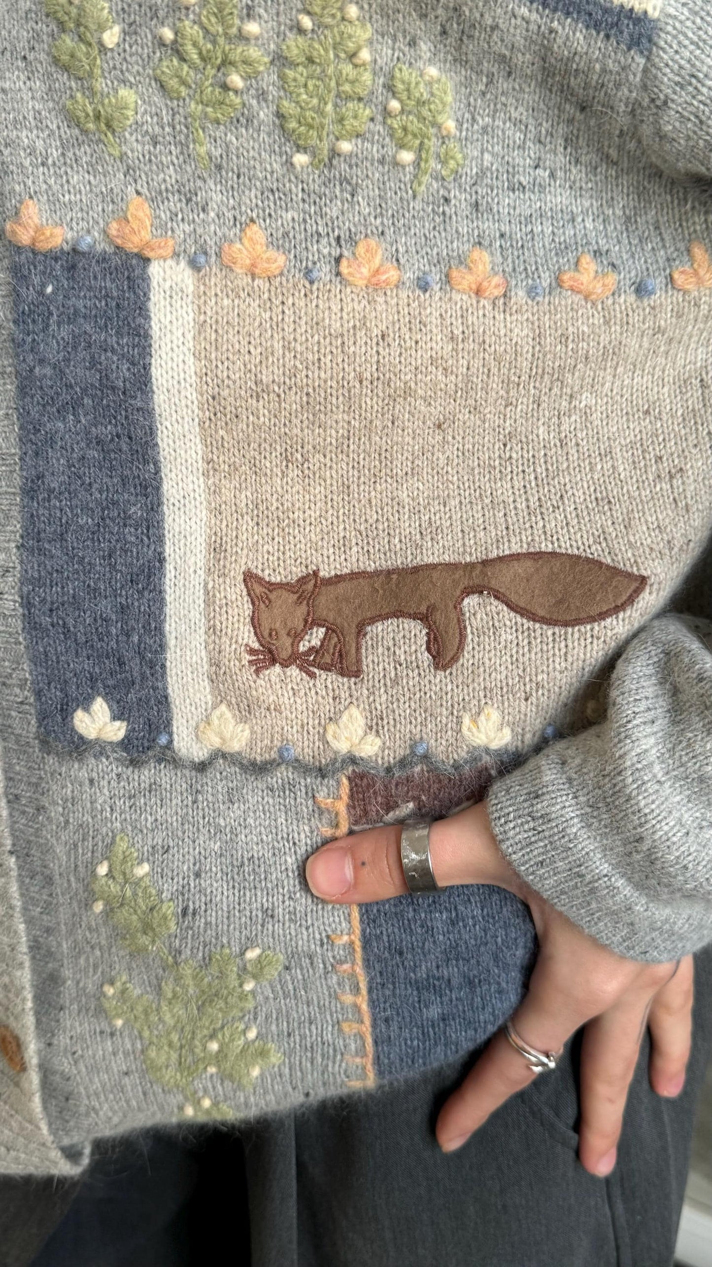 a close-up of a person's hand wearing a gray sweater with a brown fox embroidered on it.