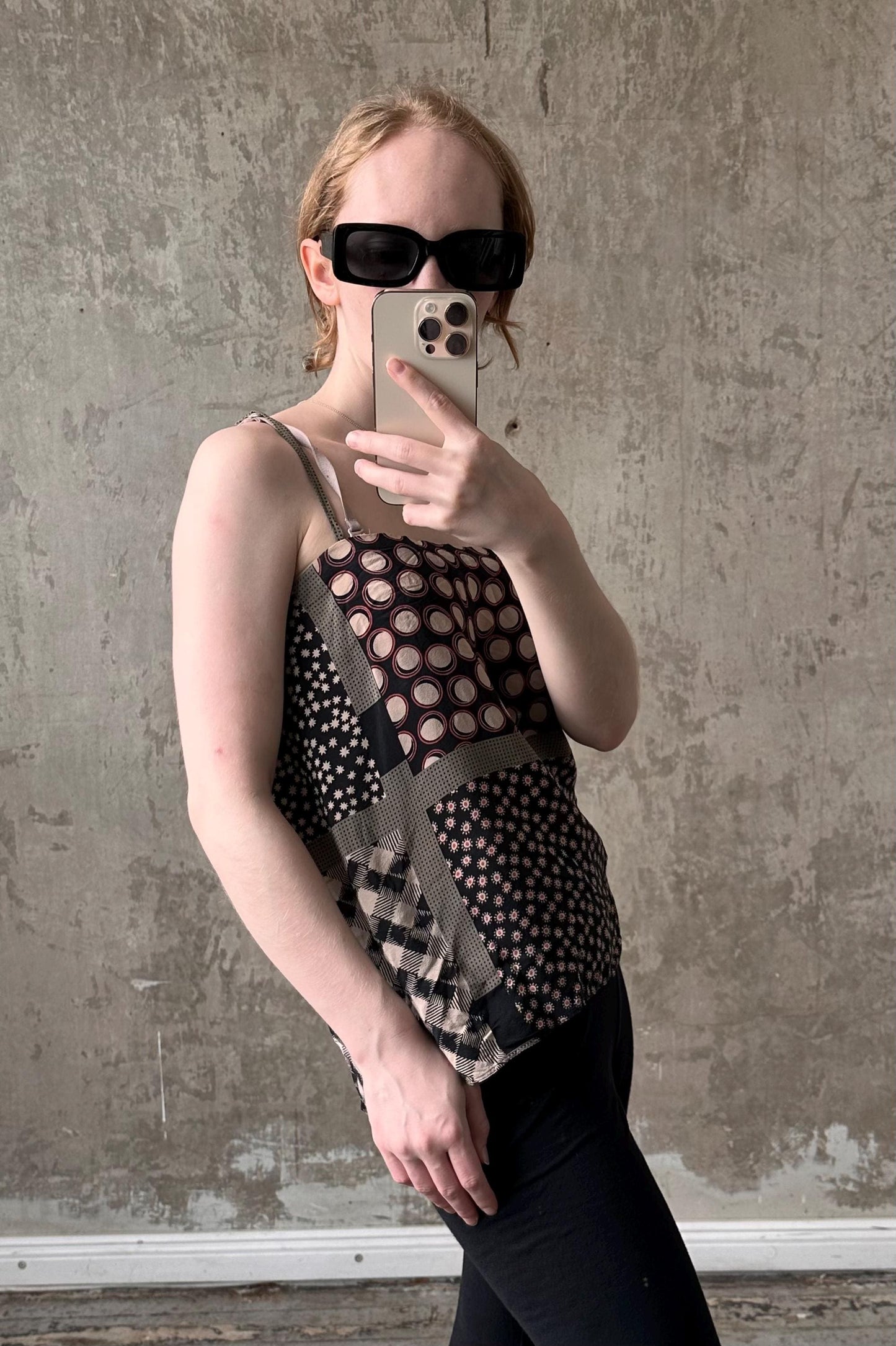 A woman wearing a patterned top and sunglasses is taking a selfie with her iPhone against a concrete wall.
