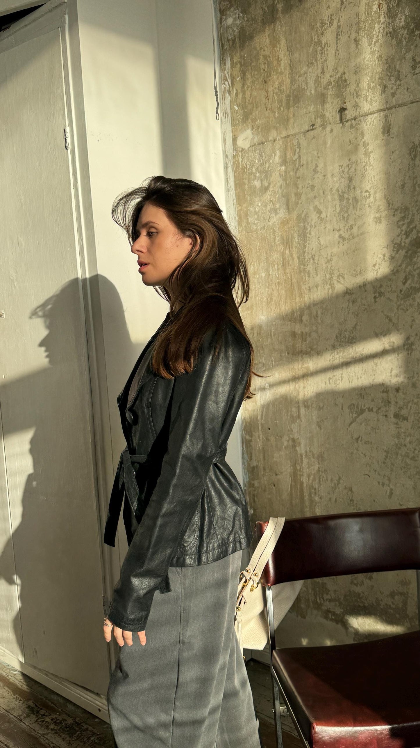 A woman in a black leather jacket stands in front of a concrete wall, with a wooden chair visible to her right.