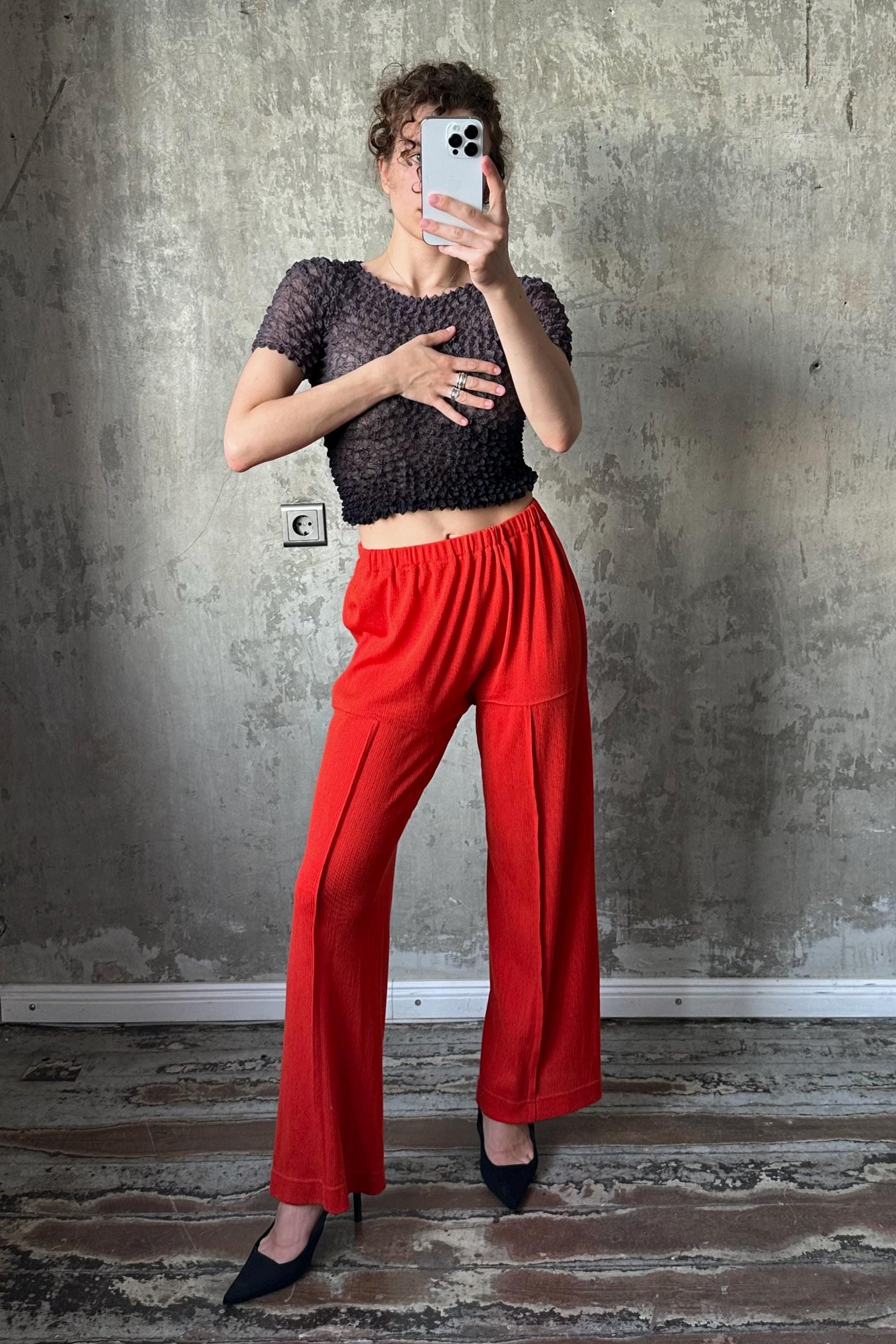 A woman is taking a selfie with a white phone, wearing a black crop top and red wide-leg pants, standing in front of a concrete wall.