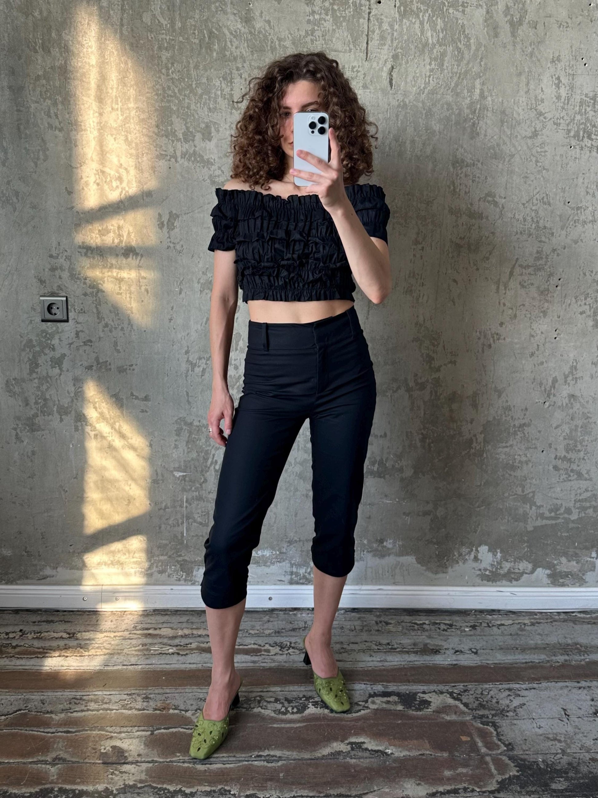 A woman with curly hair is taking a selfie with a white phone, wearing a black off-the-shoulder top and black pants, and green shoes.