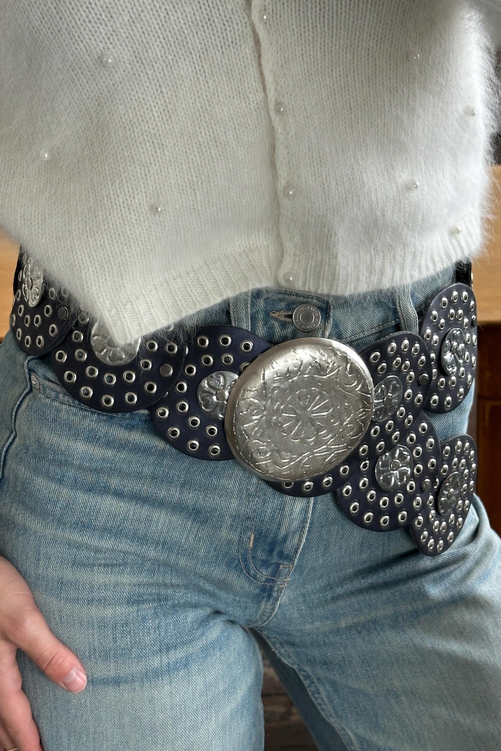 A close-up of a person wearing a white sweater and blue jeans, with a large, decorative belt featuring a metal buckle.