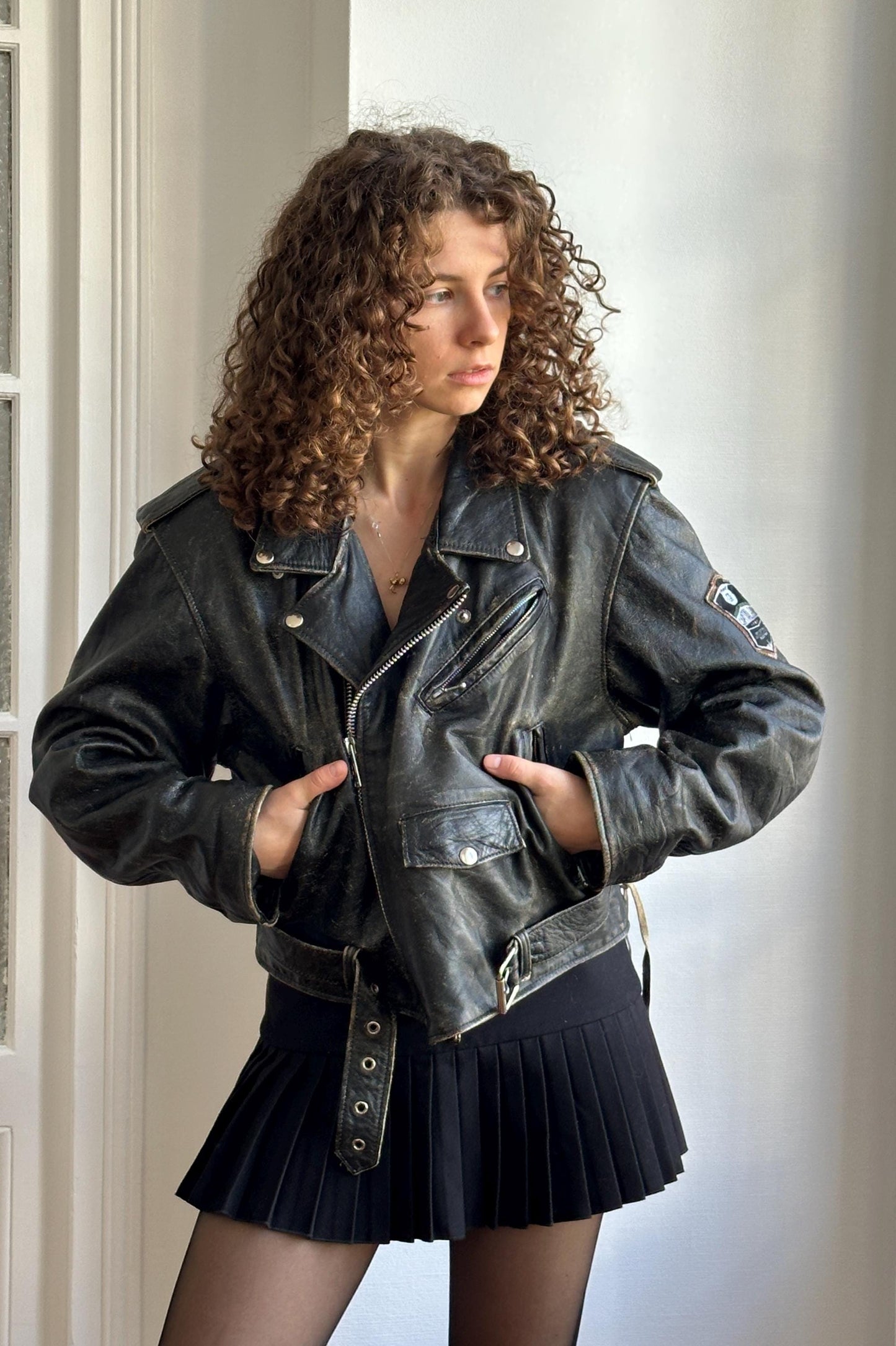 A woman with curly hair wearing a black leather jacket and a black pleated skirt stands in a room with a white wall.