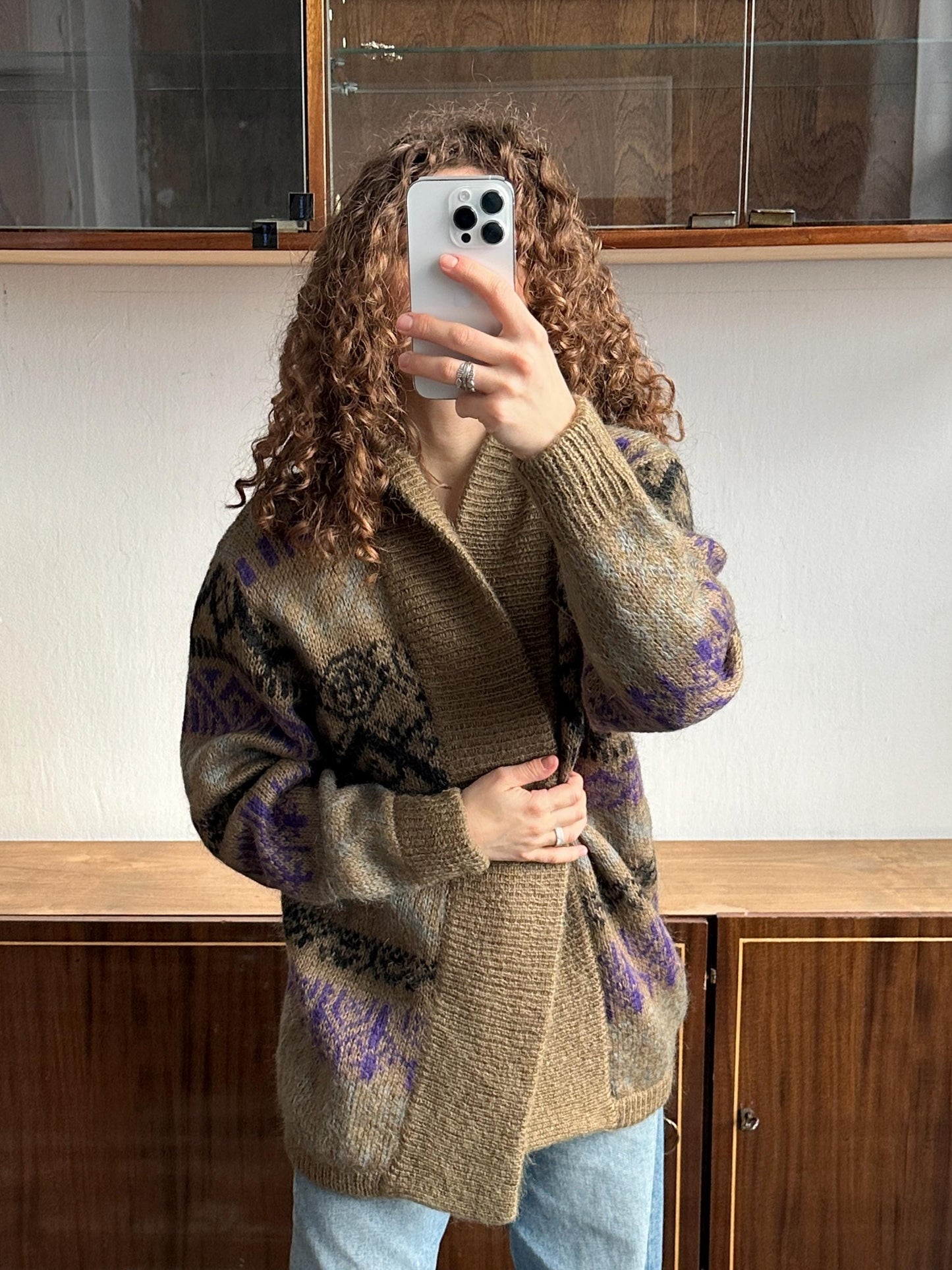 A person with curly hair is taking a selfie with a smartphone, wearing a patterned sweater.