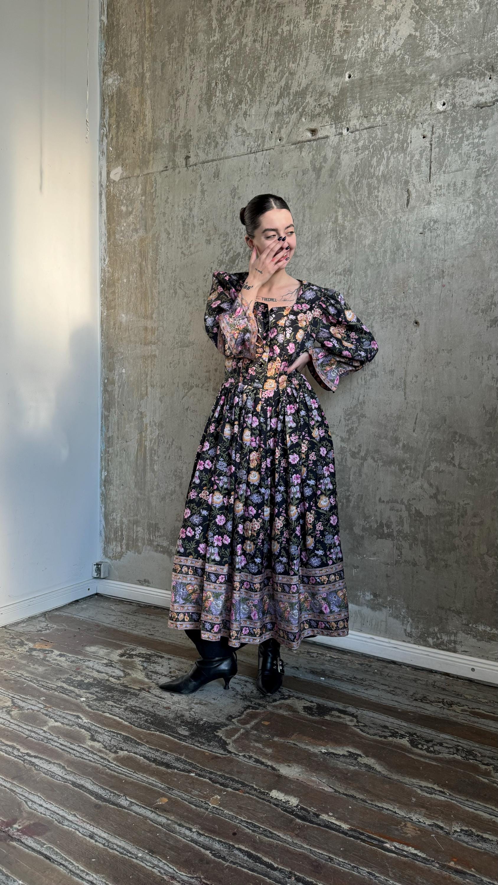 A woman wearing a long, floral-patterned dress stands in a room with a concrete wall and wooden floor.