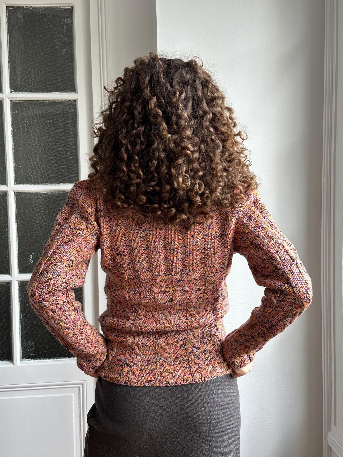 A person with curly hair is standing in front of a window, wearing a colorful sweater.