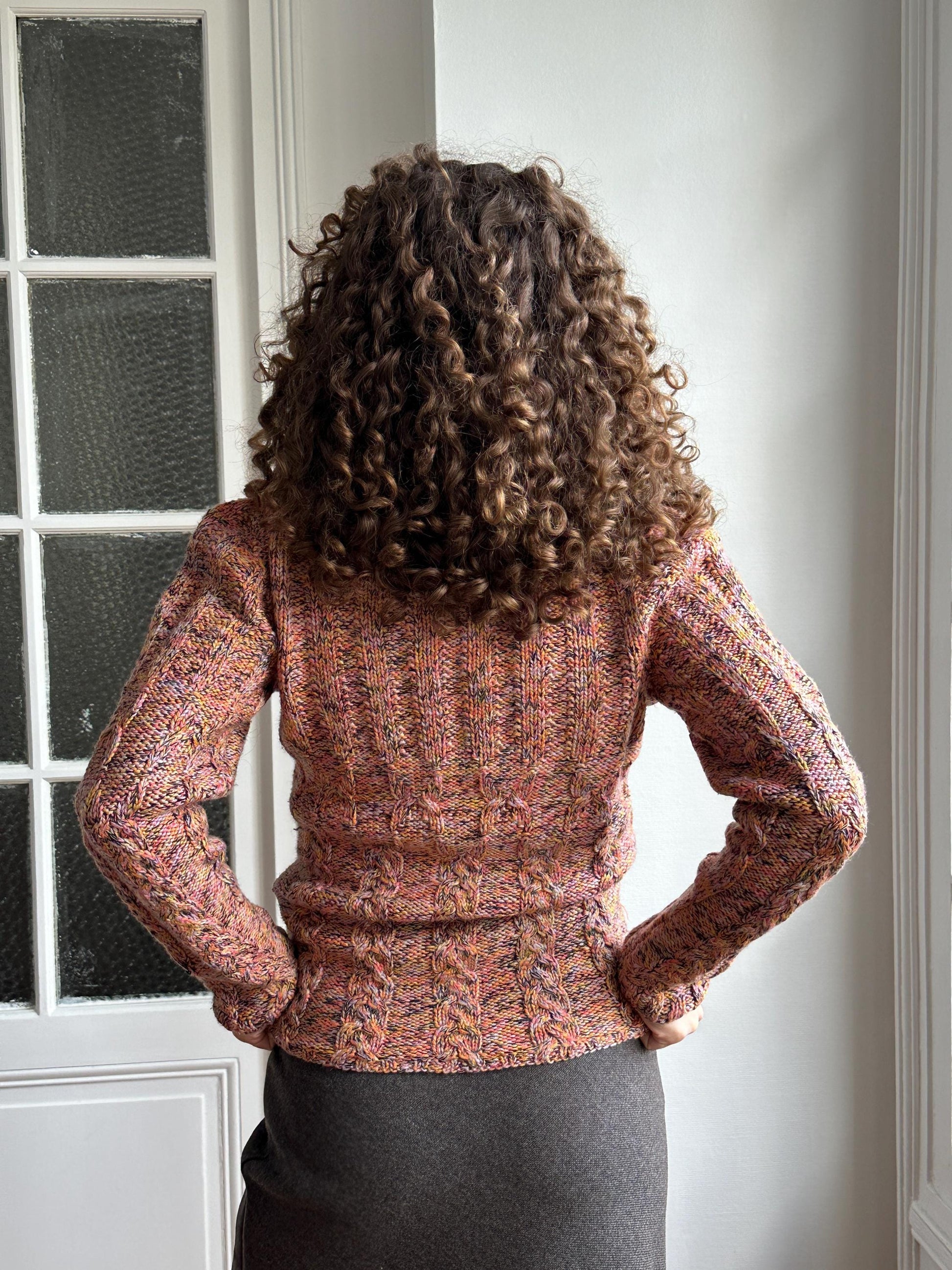 A person with curly hair is standing in front of a window, wearing a colorful sweater.