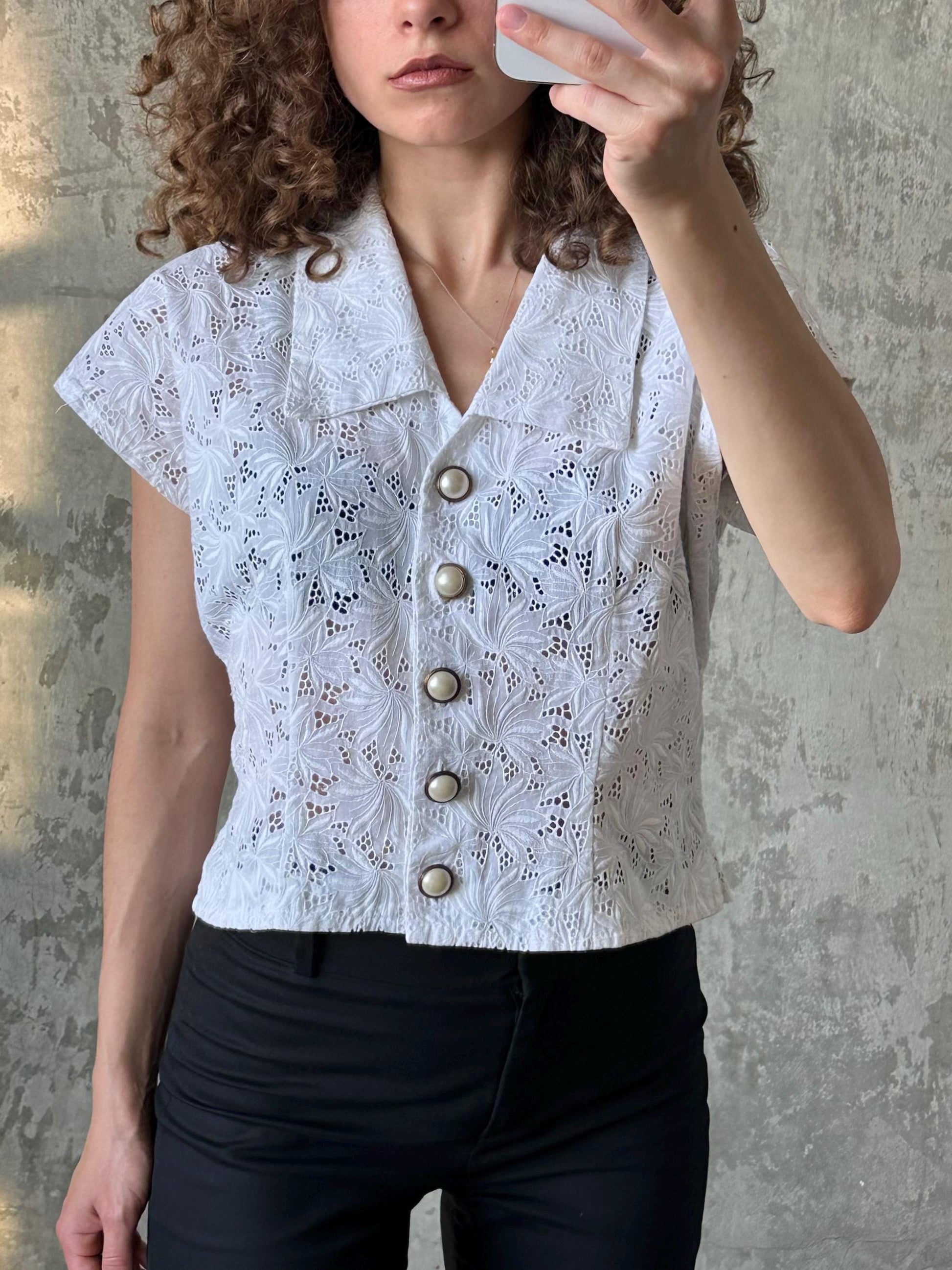 A woman with curly hair is taking a selfie with a white lace top and black pants.