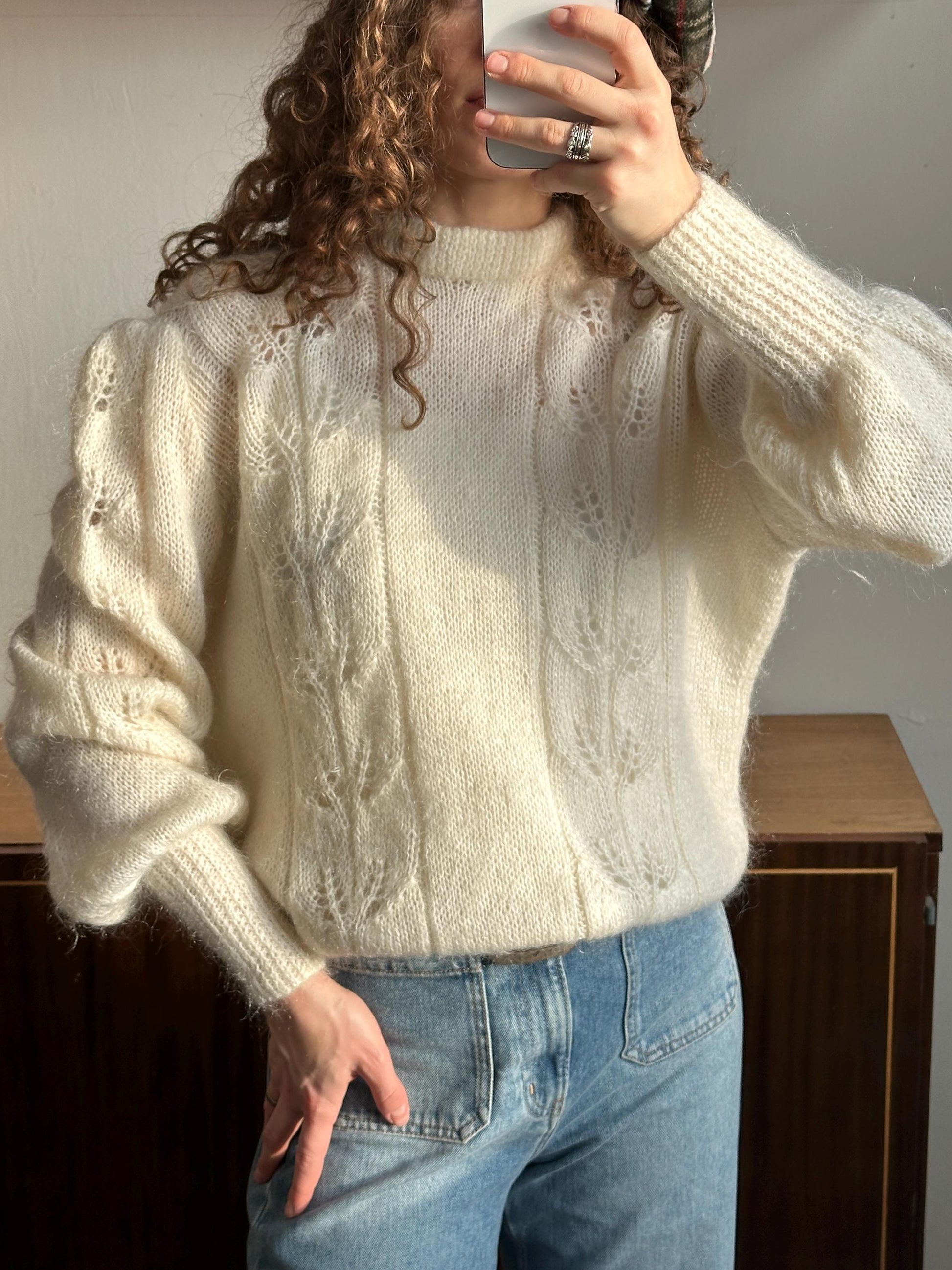 A person is taking a selfie with a white sweater featuring lace-like patterns, and they are holding a phone up to their face.