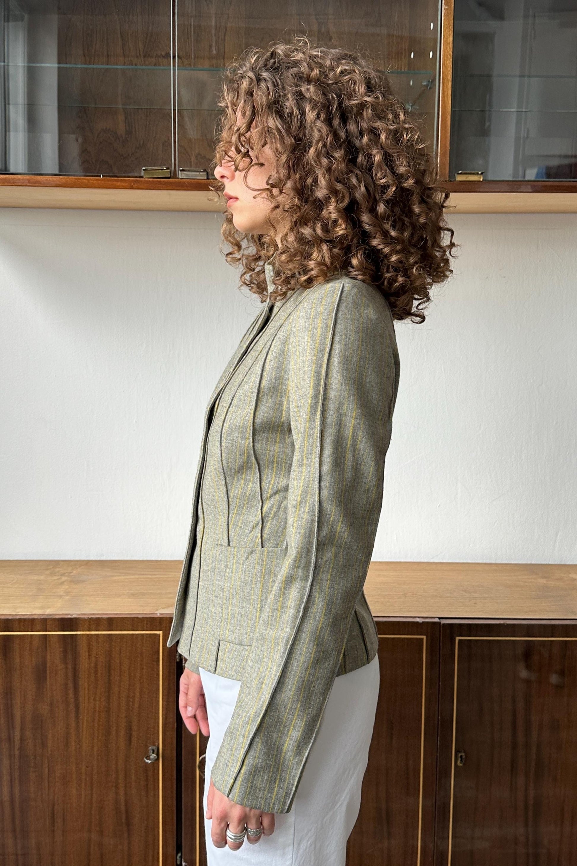 A woman with curly hair wearing a striped jacket stands in front of a wooden cabinet.