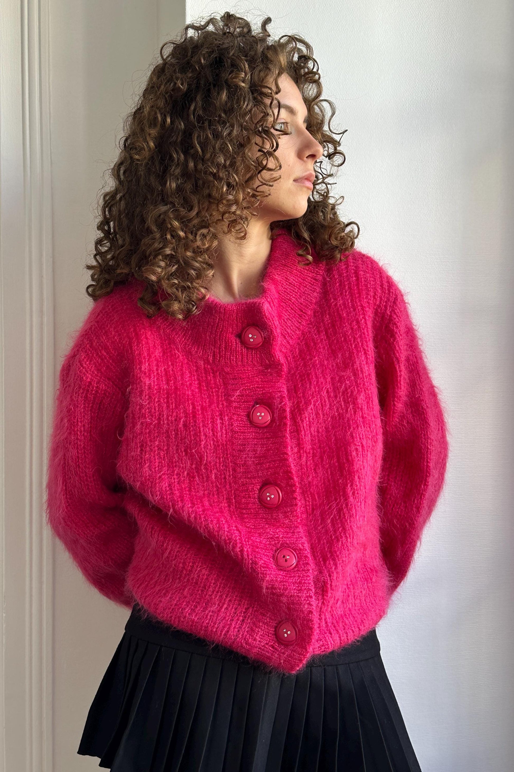 A woman with curly hair wearing a bright pink cardigan and a black skirt stands in front of a white wall, looking to her left.