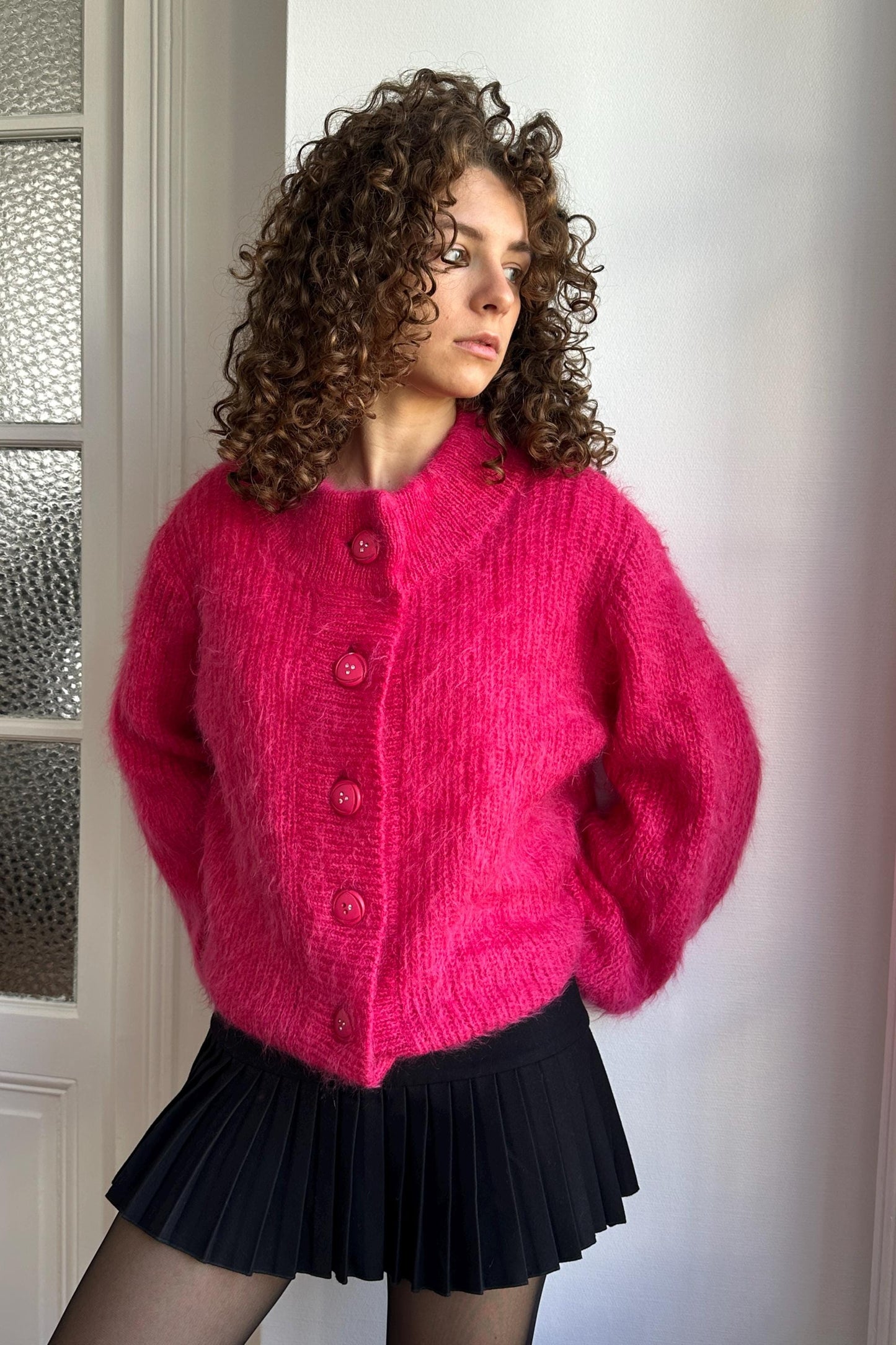 A woman with curly hair wearing a pink cardigan and a black pleated skirt stands in front of a white wall.