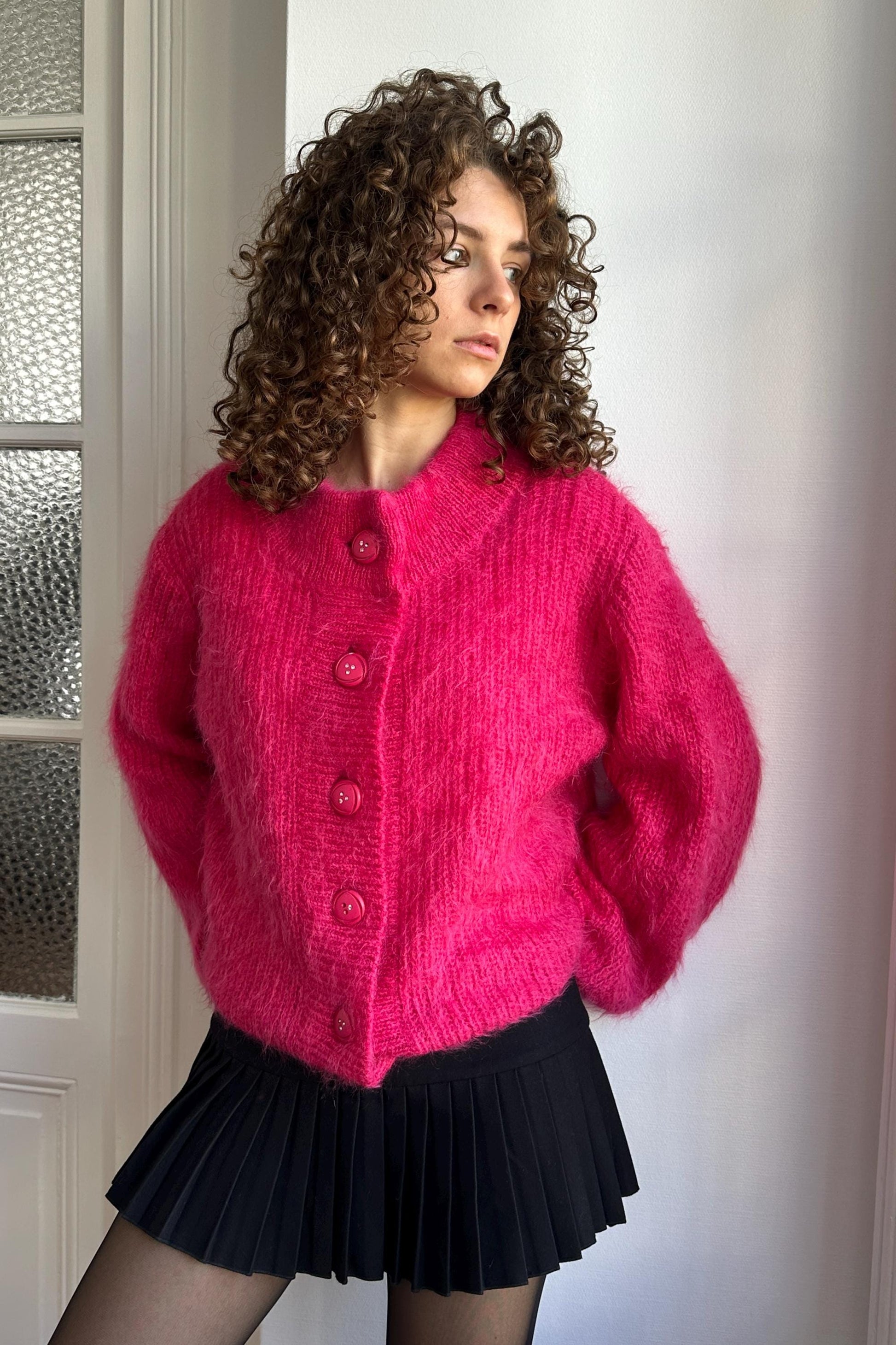 A woman with curly hair wearing a pink cardigan and a black pleated skirt stands in front of a white wall.