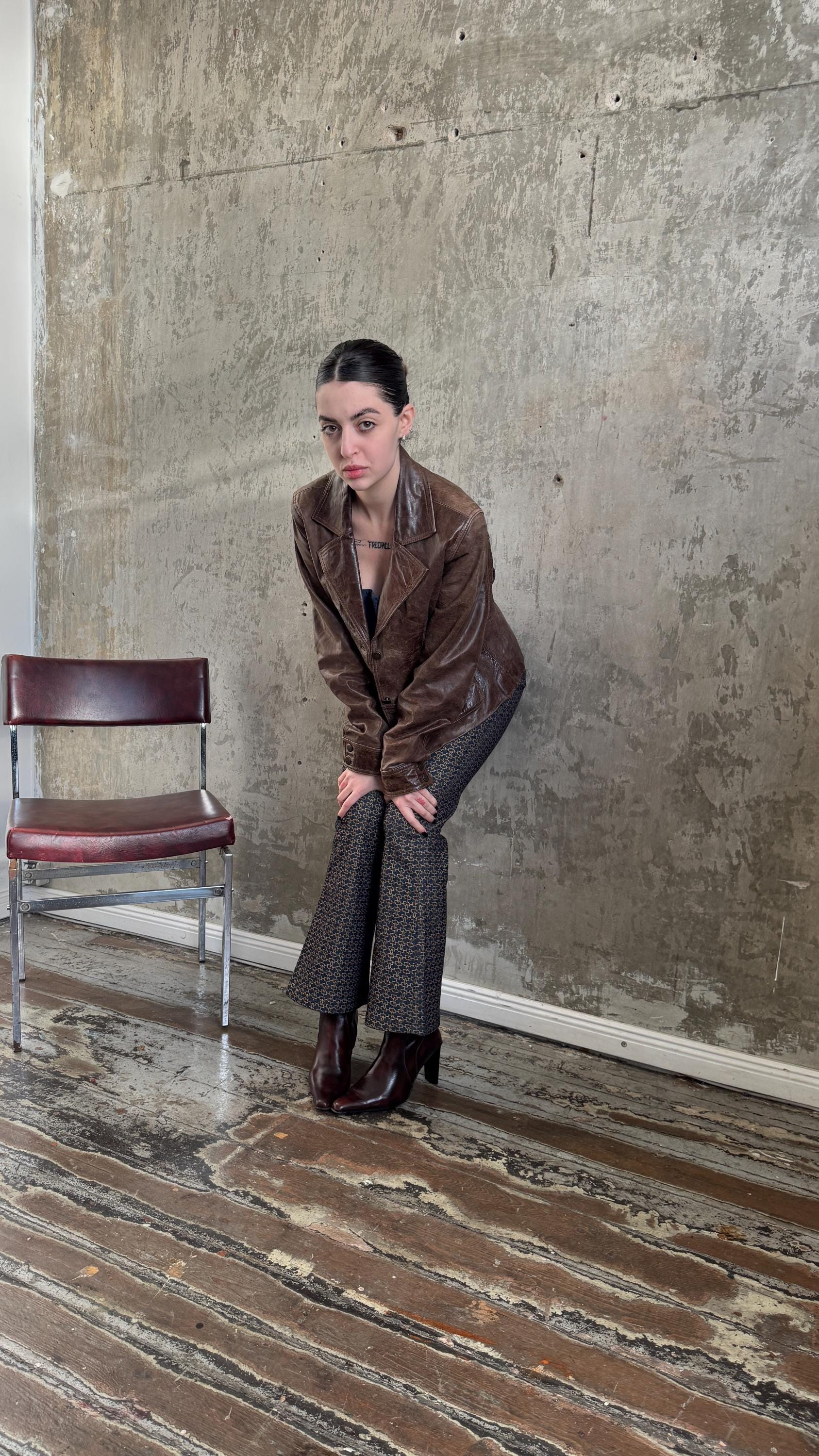 A woman in a brown leather jacket and patterned pants is posing in front of a concrete wall, leaning against it with her hands on her knees.