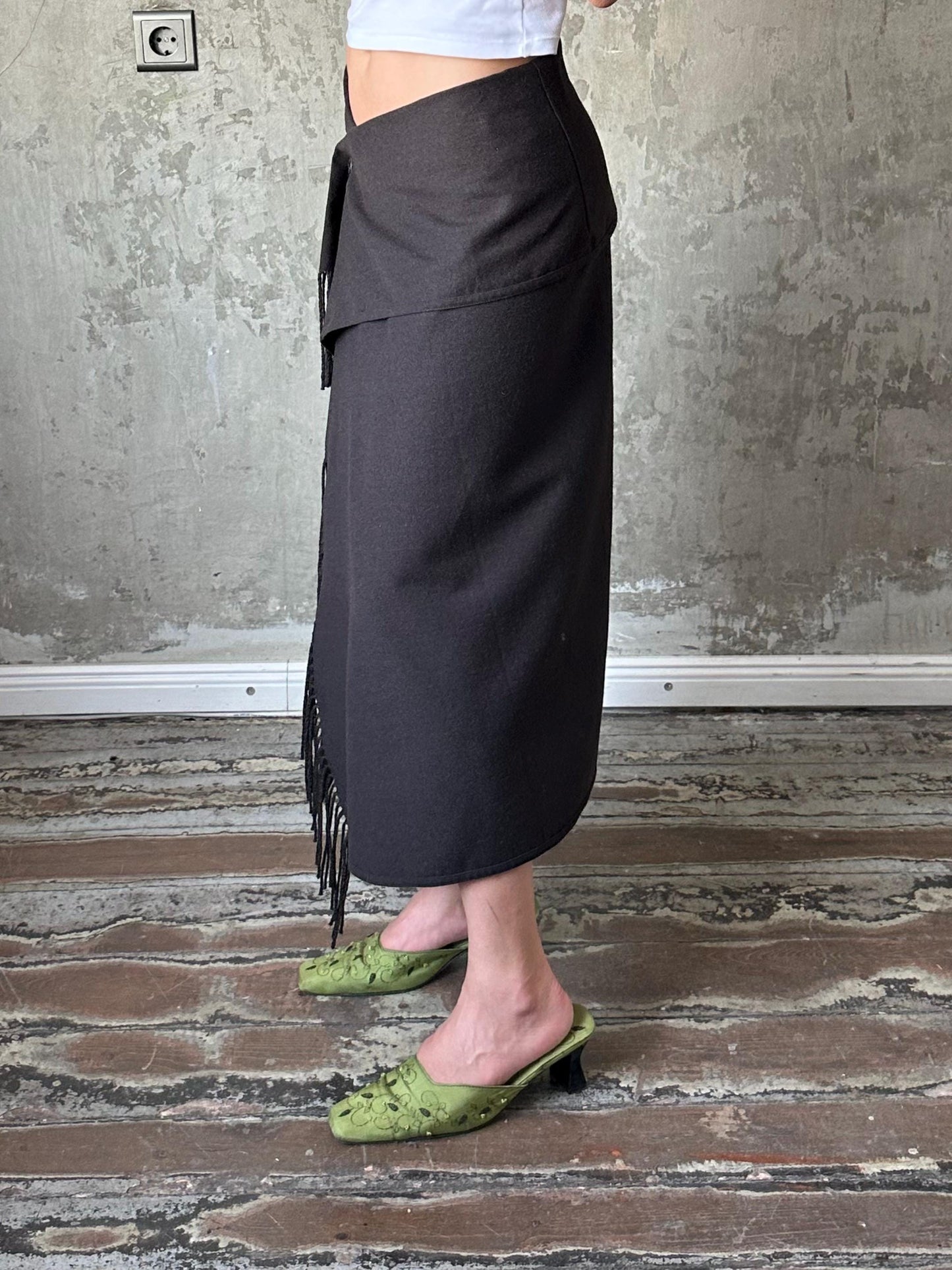 A person wearing a black skirt and green shoes stands in front of a concrete wall.