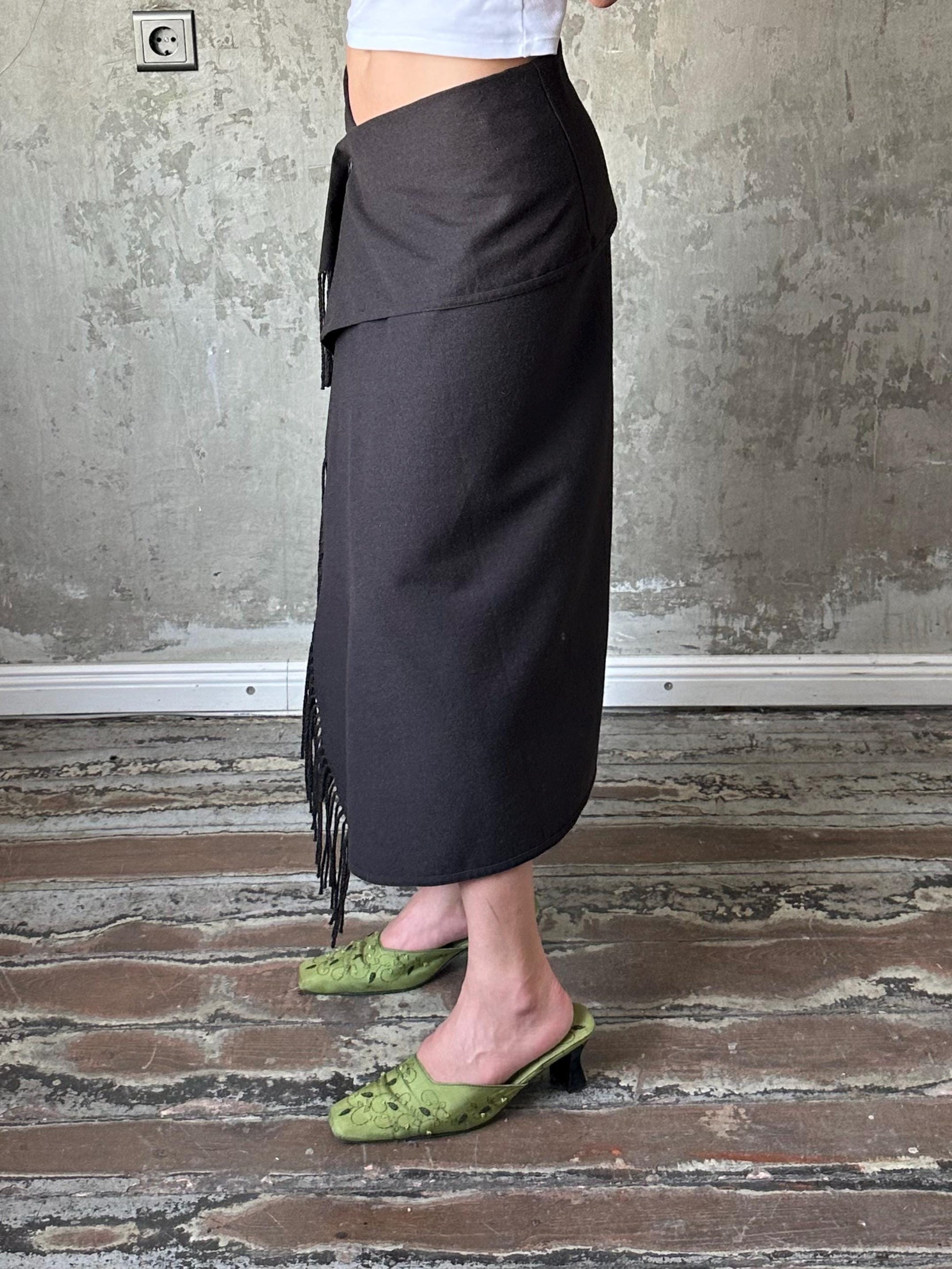 A person wearing a black skirt and green shoes stands in front of a concrete wall.
