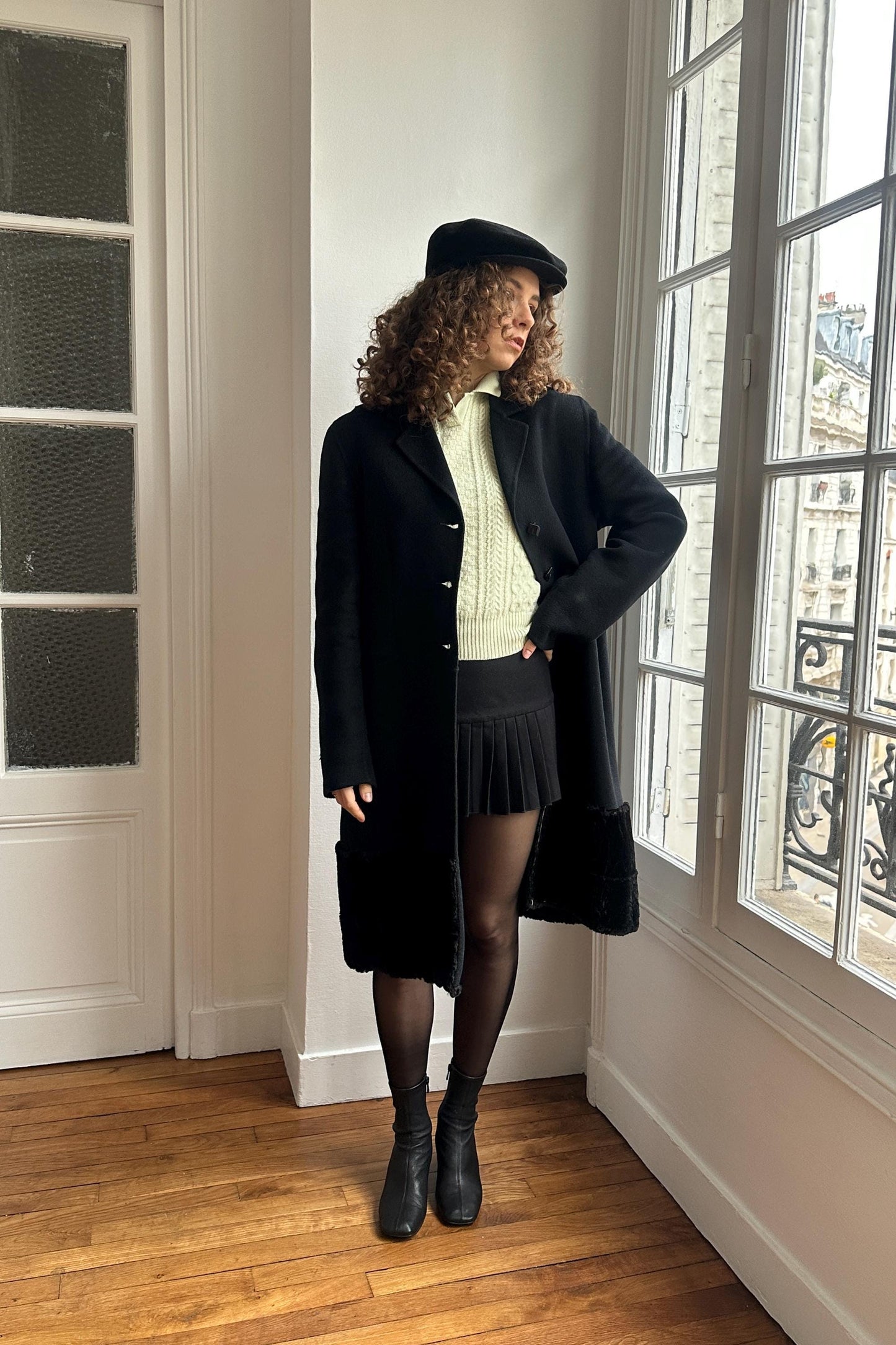 A woman in a black coat, hat, and skirt stands in a room with a large window, looking out at the view.
