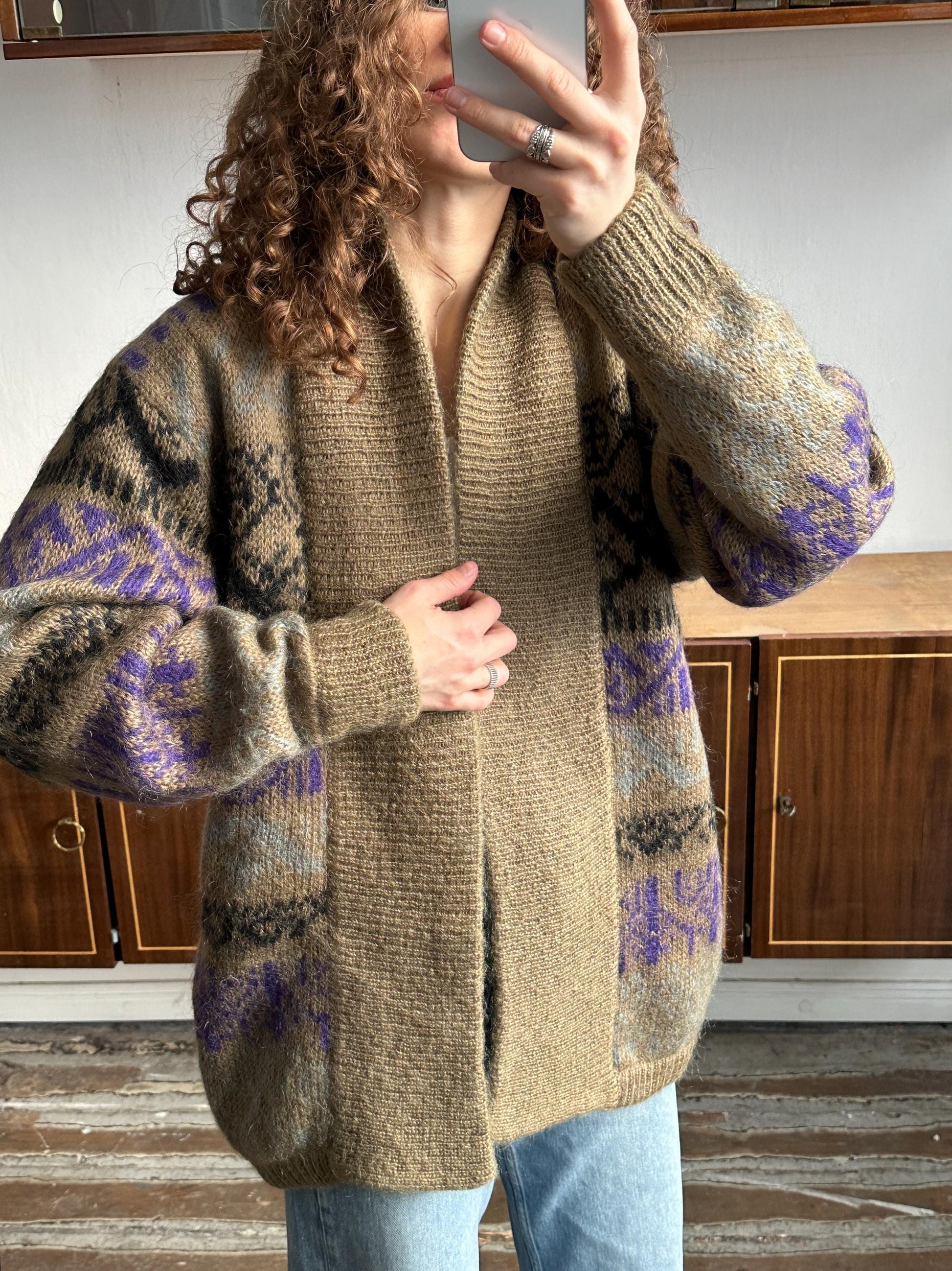 A person with curly hair is taking a selfie with a smartphone, wearing a beige cardigan with purple and black patterned designs.