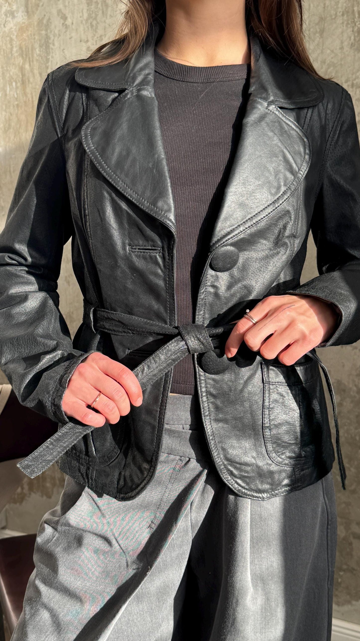 A person wearing a black leather jacket and gray pants, with their hands adjusting the belt.