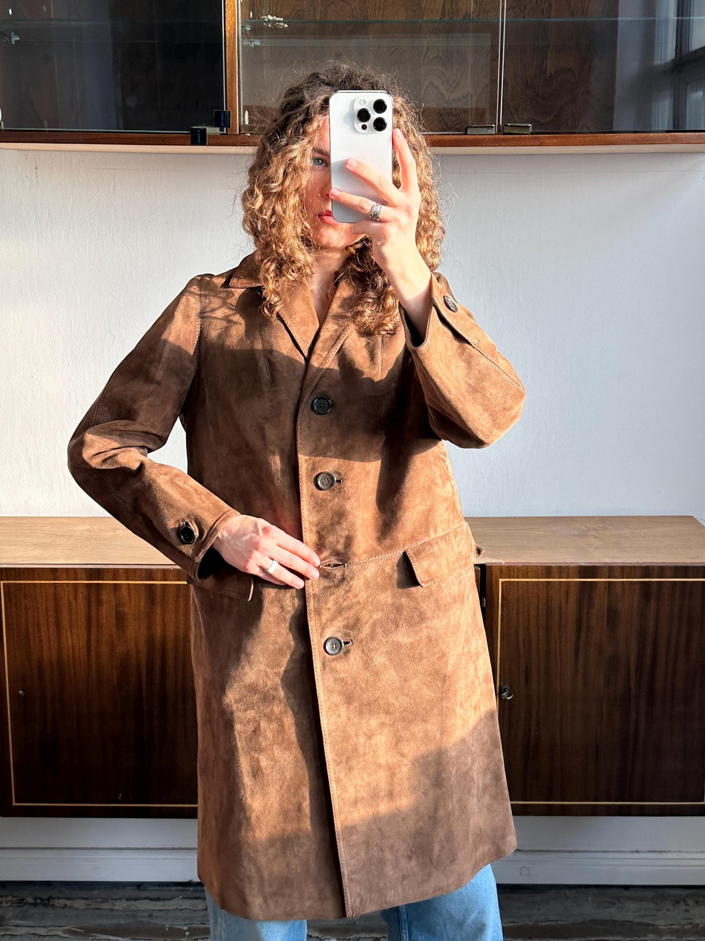 A person with curly hair is taking a selfie with a white phone, wearing a brown coat.