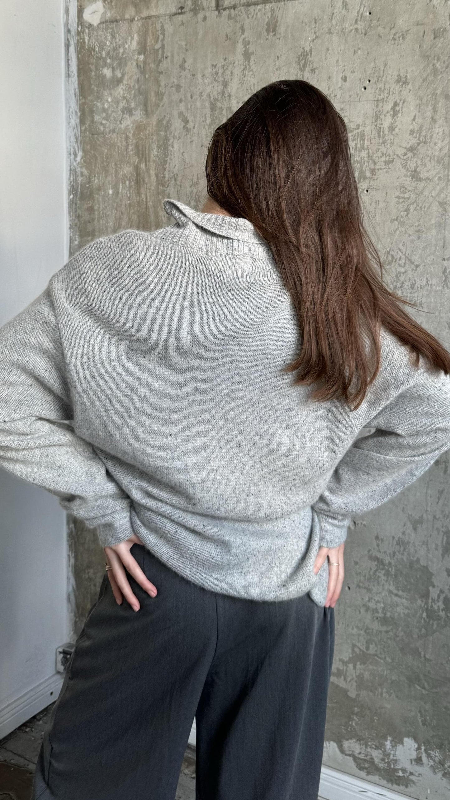 A person with long brown hair is standing in front of a concrete wall, wearing a gray sweater and dark pants.