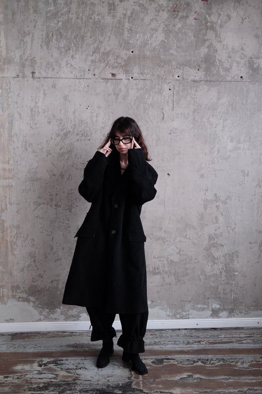 A person wearing a long black coat stands in a room with a concrete wall.