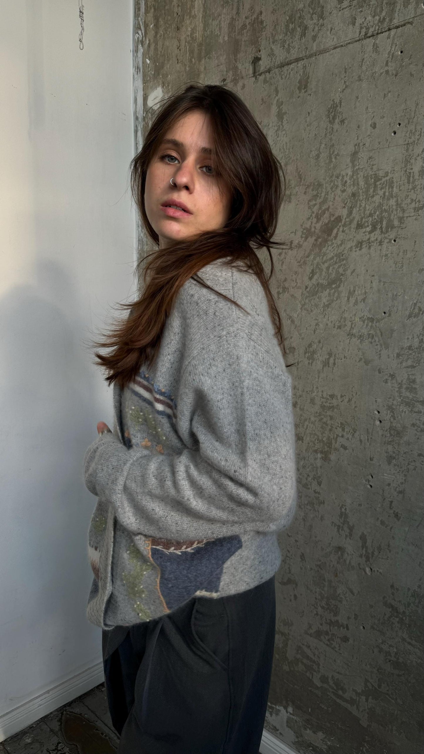 A young woman with long brown hair stands in front of a concrete wall, wearing a gray sweater with a colorful design.