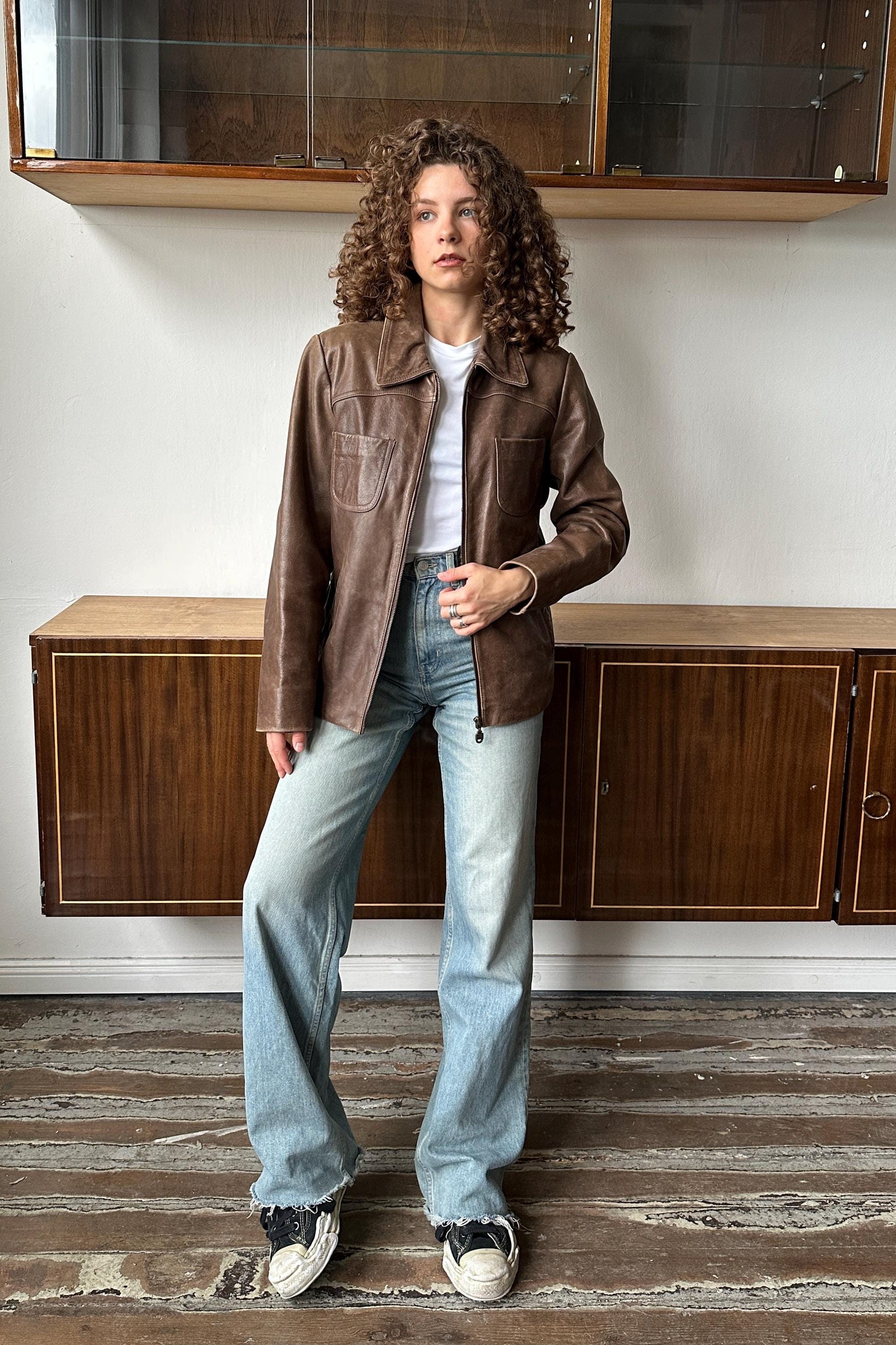A woman with curly hair wearing a brown leather jacket, white shirt, and blue jeans stands in a room with wooden cabinets.