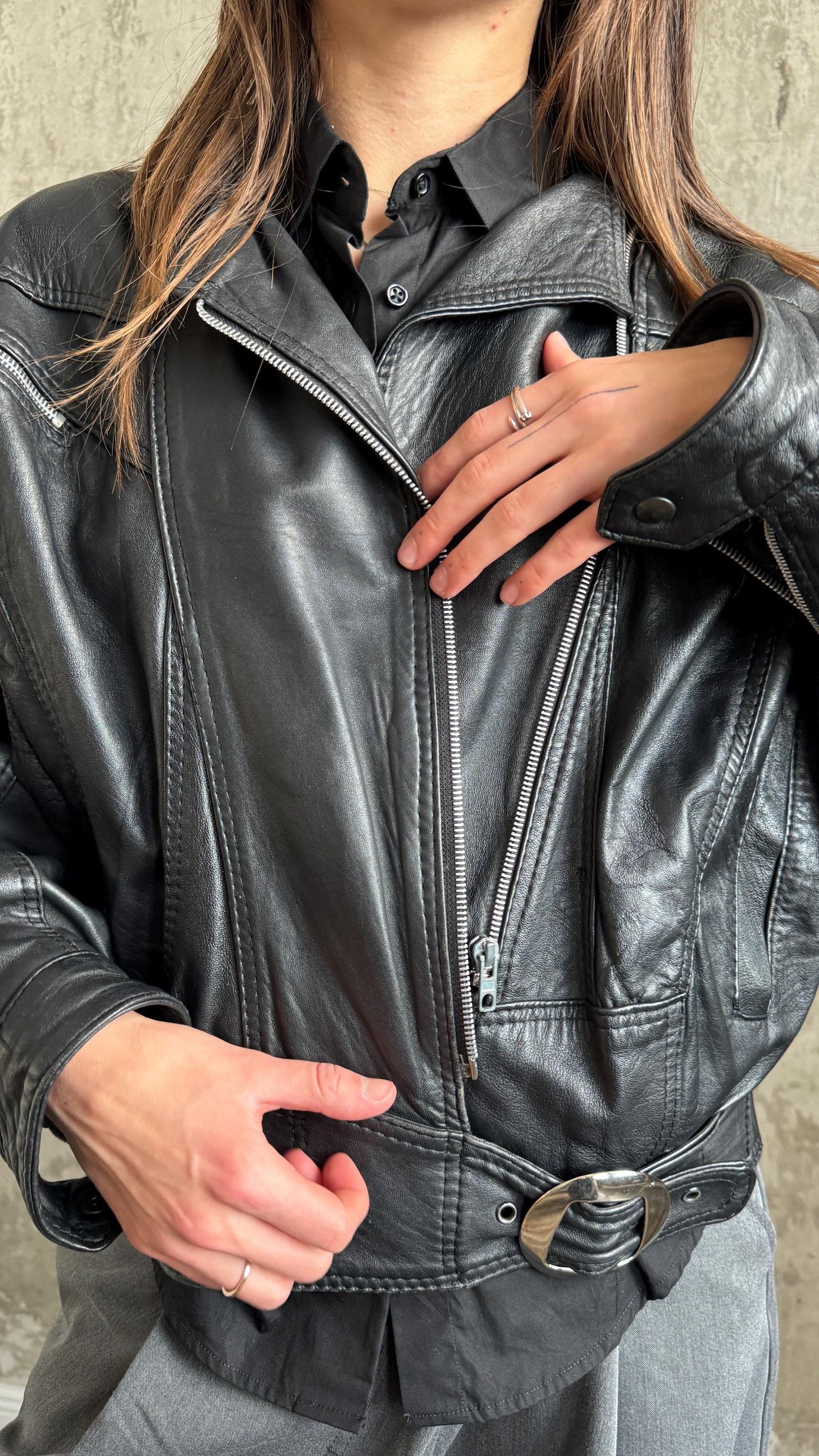 A person wearing a black leather jacket with a silver buckle, posing with their hands on their hips.