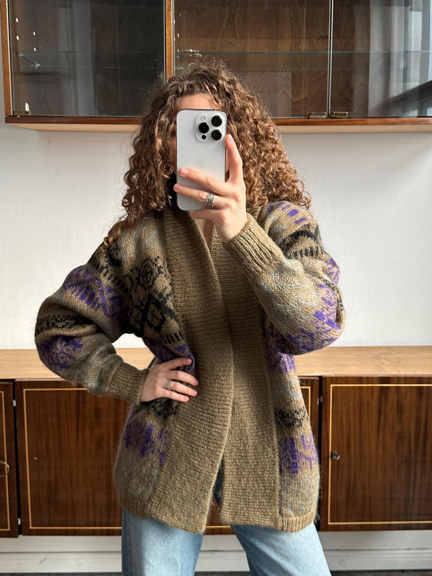 A woman with curly hair is taking a selfie with a smartphone, wearing a brown cardigan with purple and black patterns.