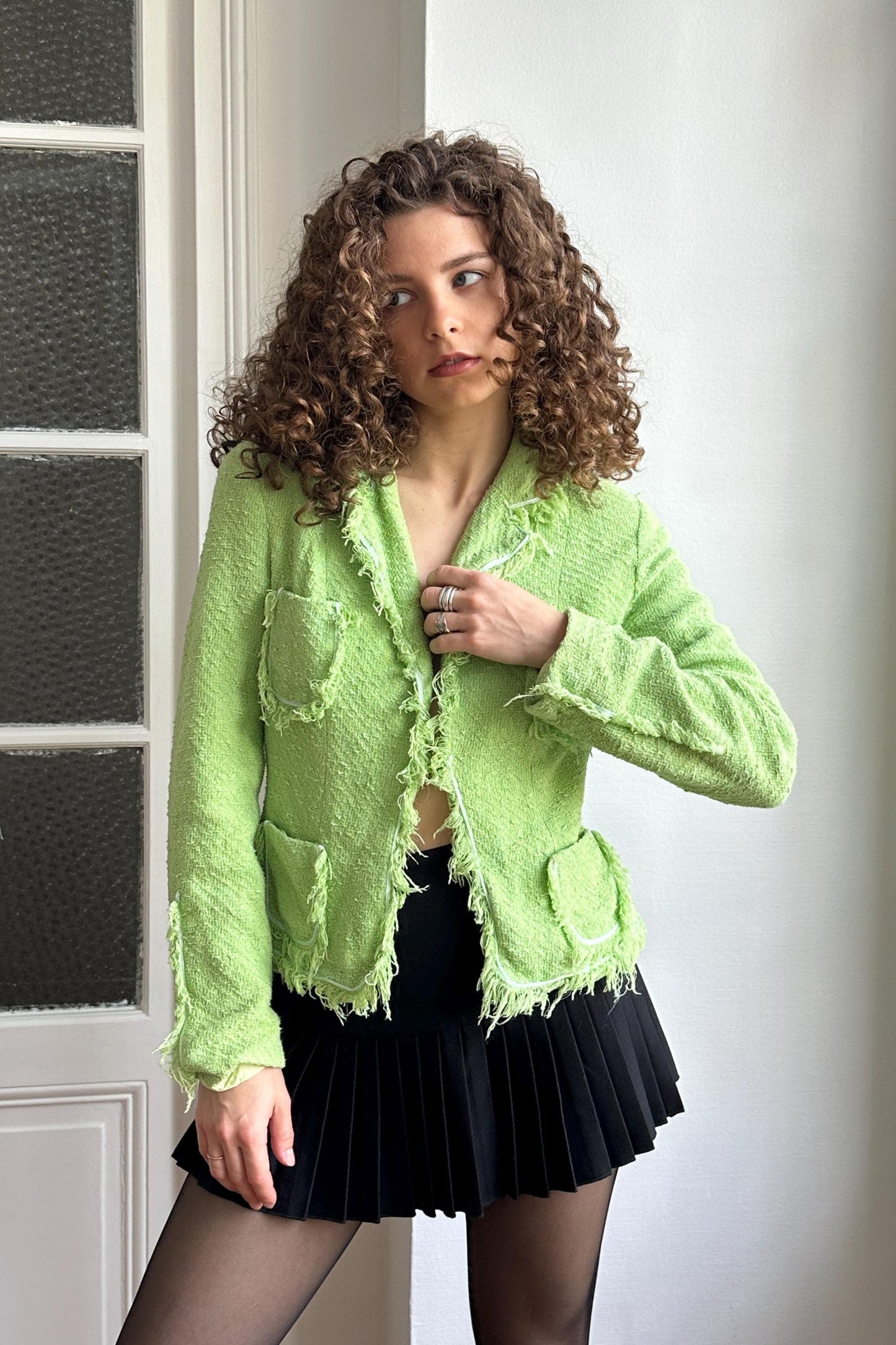 A woman with curly hair wearing a lime green jacket and a black skirt stands in front of a white door.