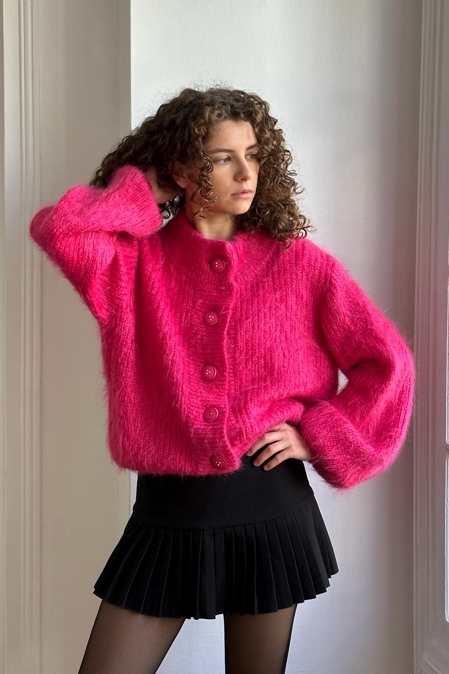 A woman with curly hair wearing a bright pink cardigan and a black pleated skirt stands in front of a white wall.