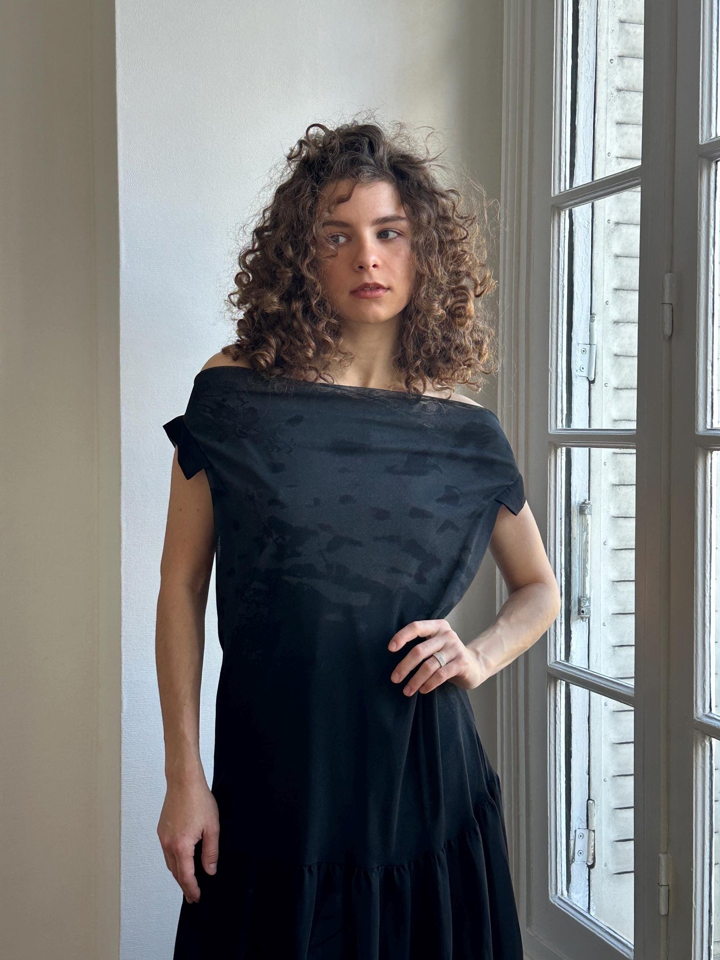 A woman with curly hair wearing a black off-the-shoulder dress stands in front of a window, looking to the side with a serious expression.