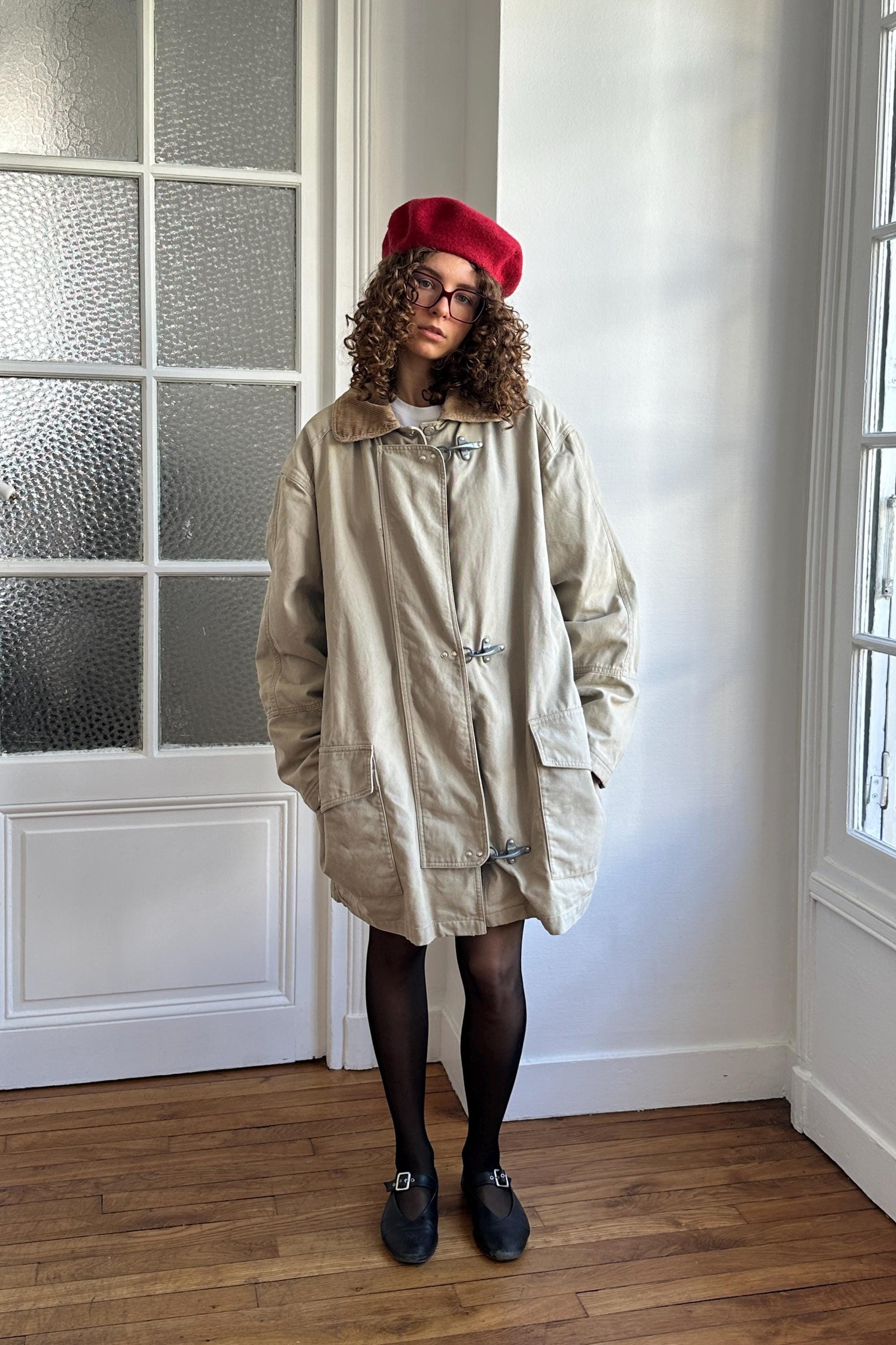 A woman wearing a beige coat, black tights, and black shoes stands in a room with a window.